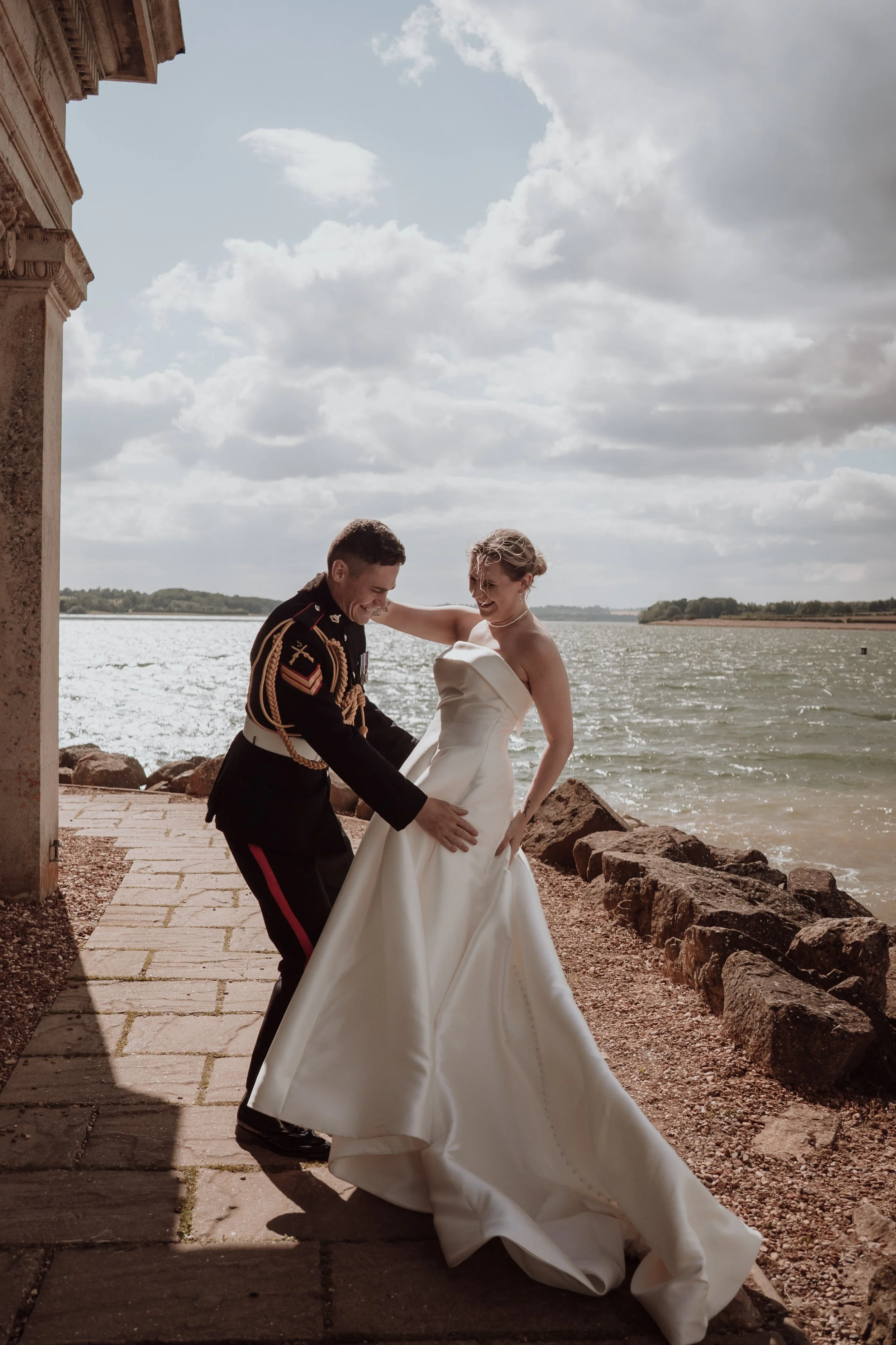 A bride in a white wedding dress and a groom in a military uniform dancing by a lake, with rocks and a cloudy sky in the background.