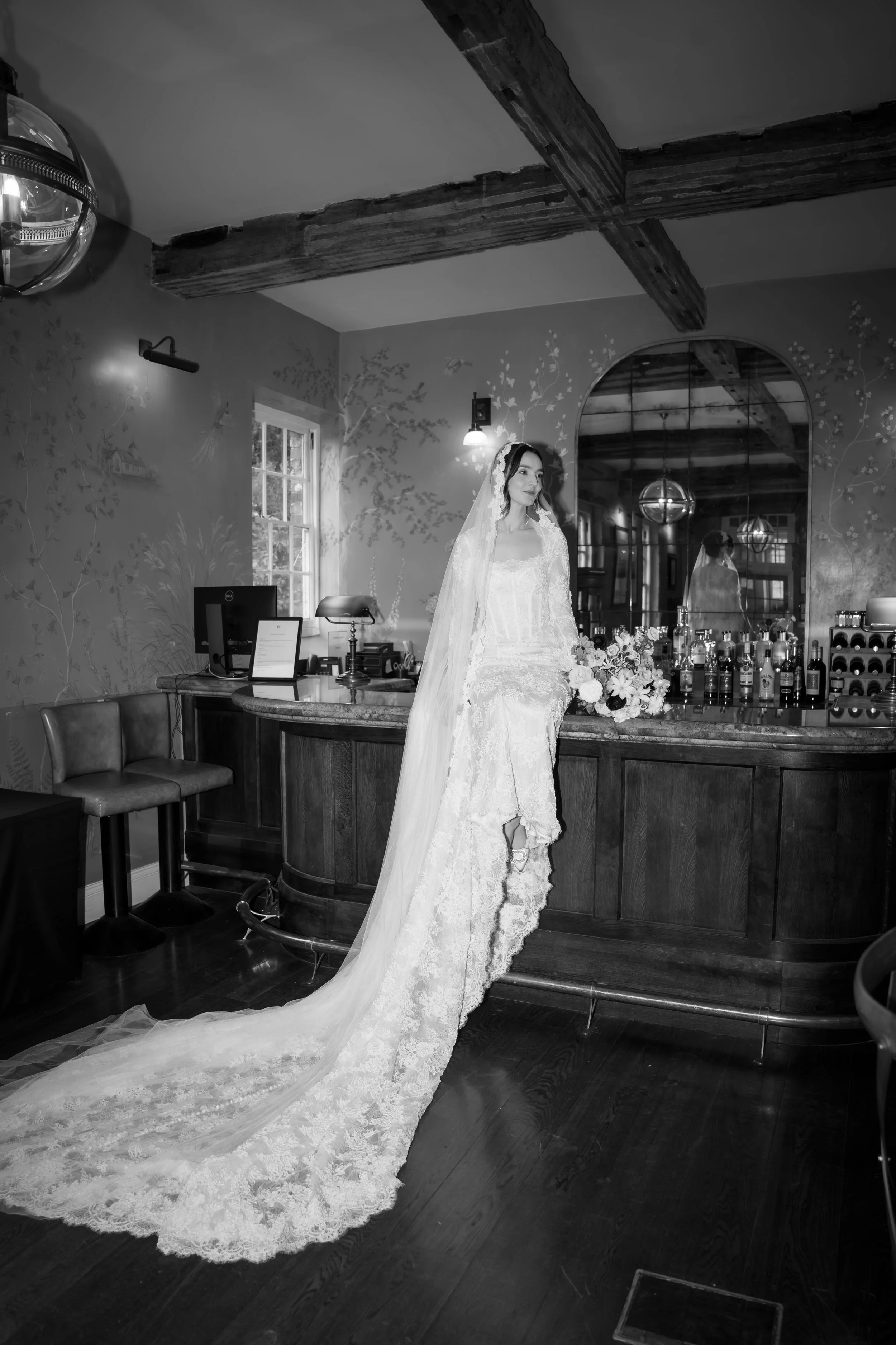 Bride in wedding dress and veil standing next to a bar in a decorated room with floral wallpaper, wooden ceiling beams, and a large mirror reflecting the bride.