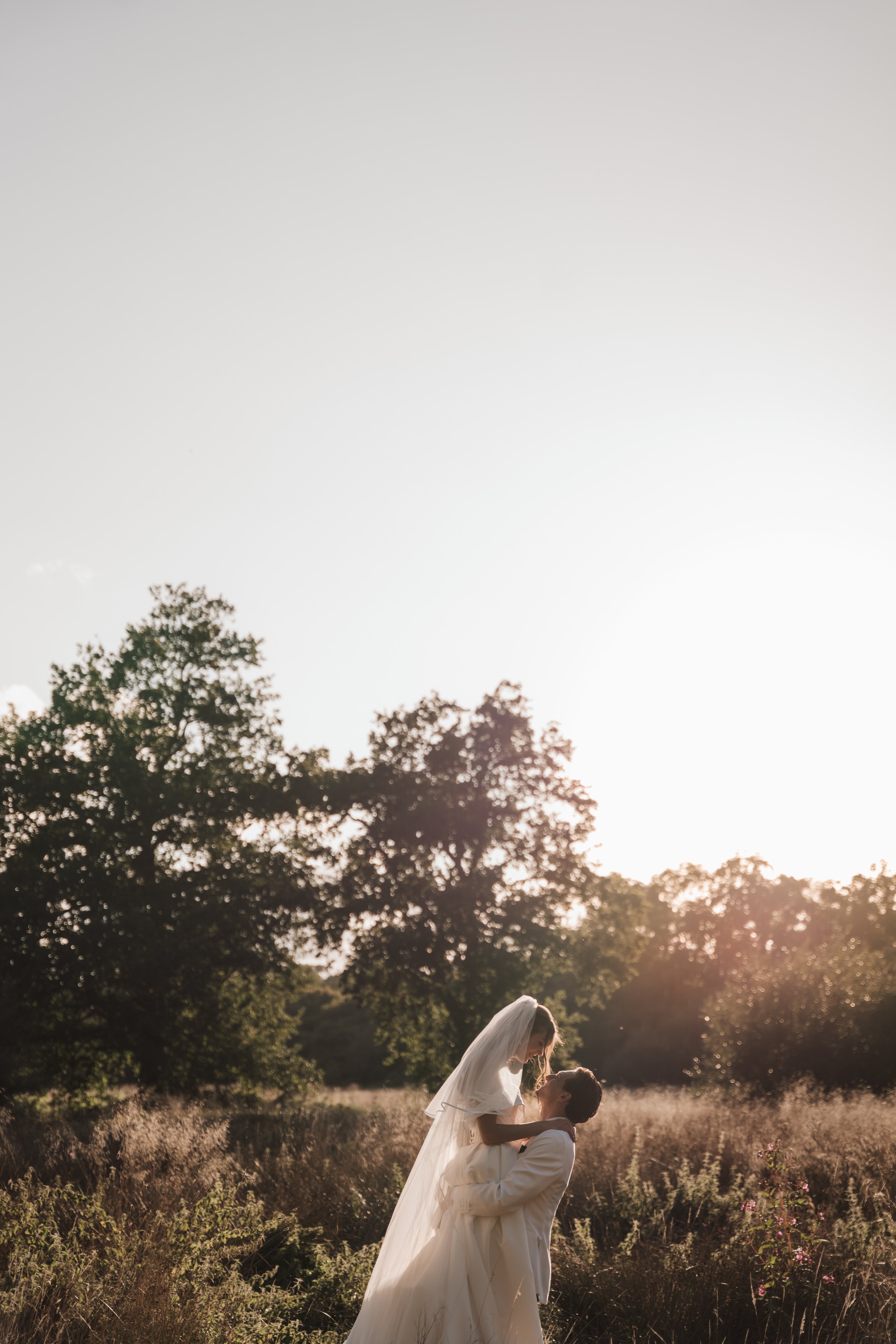 A bride and groom sharing a romantic moment outdoors during sunset in a field surrounded by trees.