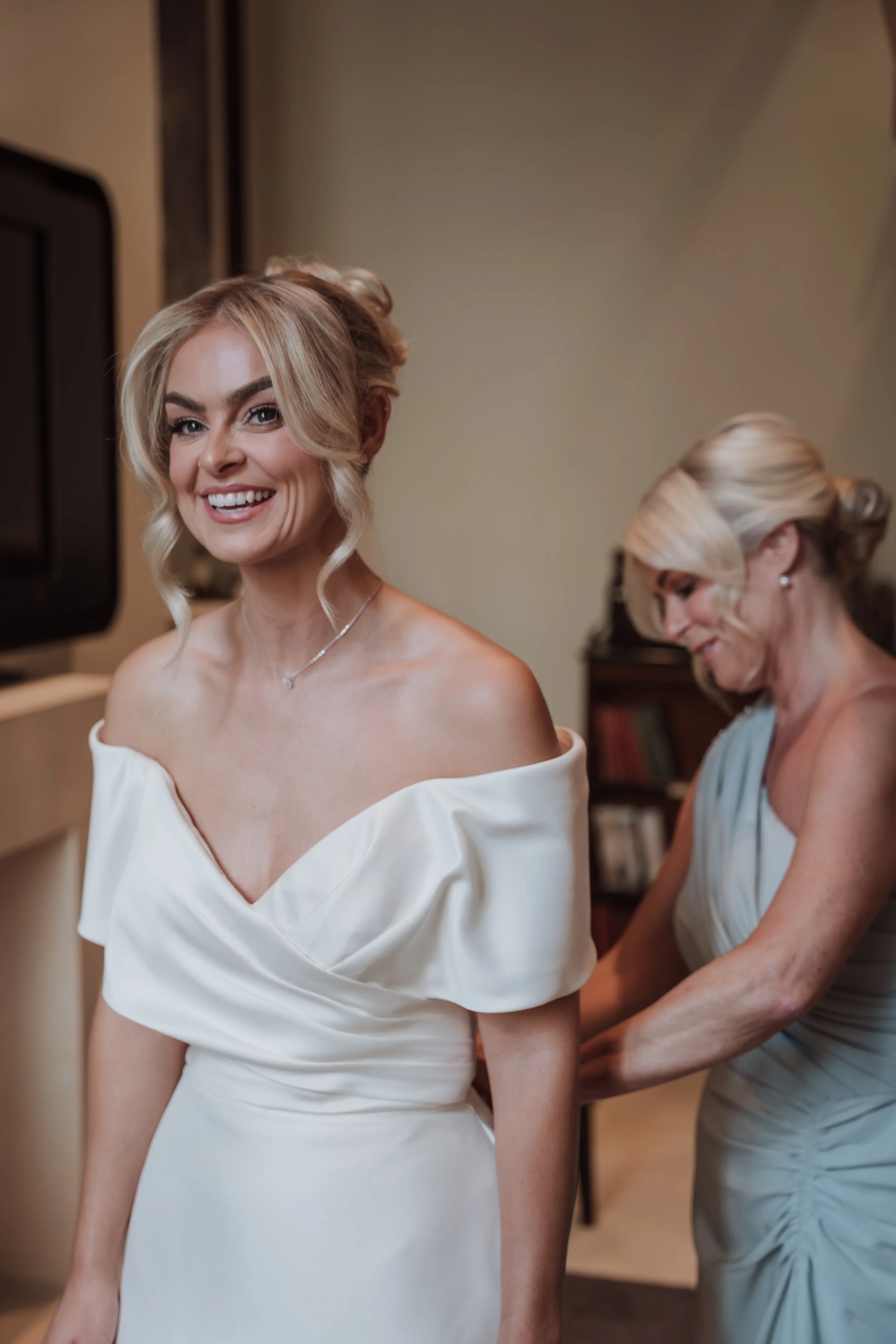 A smiling woman in a white off-the-shoulder dress getting ready, with another woman assisting her in a light blue dress in the background, in a room with a television and a bookshelf.