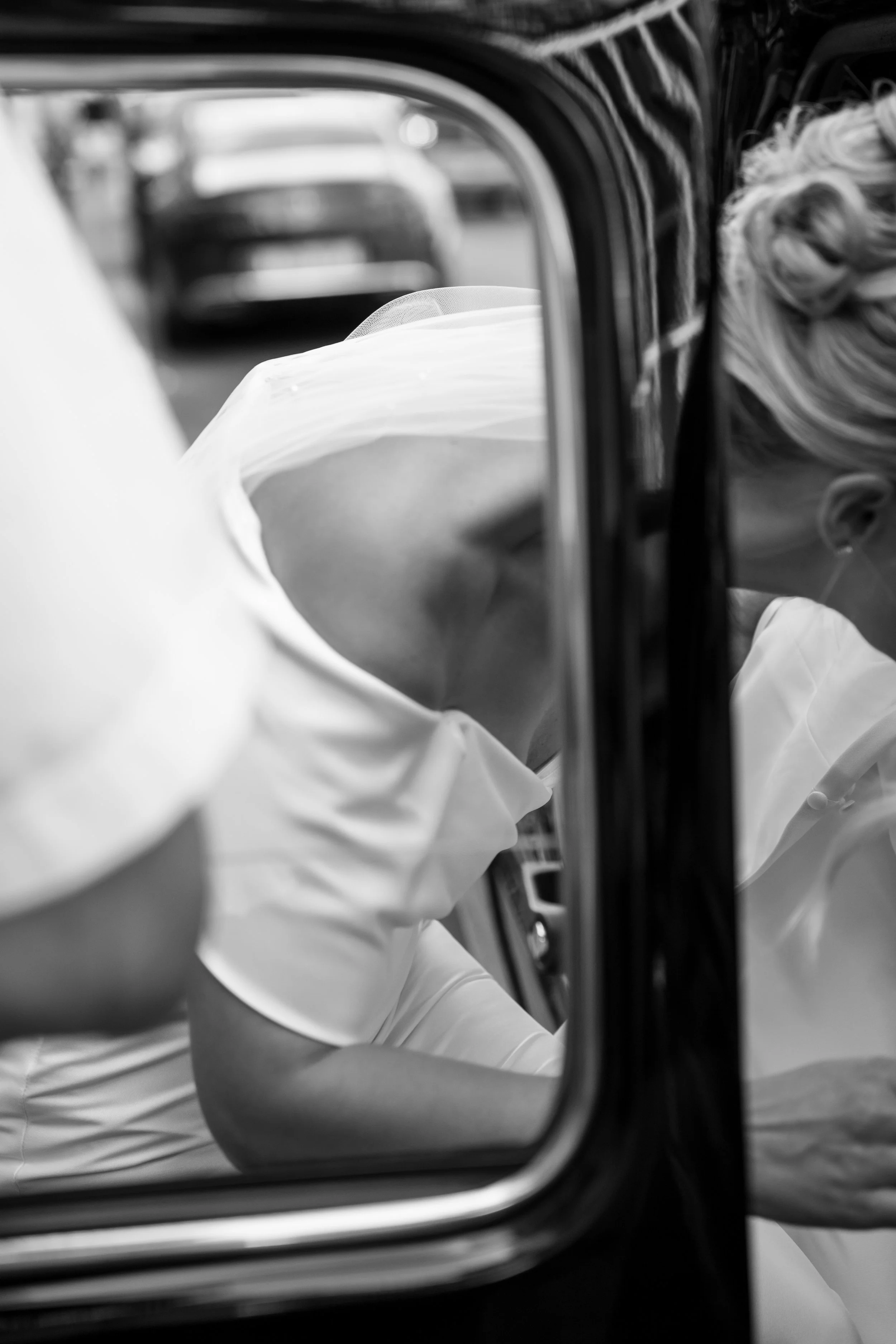 Black and white photo of a person wearing a white shirt taking a selfie through a round mirror, with their head resting on their arm. The person's face is obscured, and their long, wavy hair is visible. The reflection captures a distorted view of the