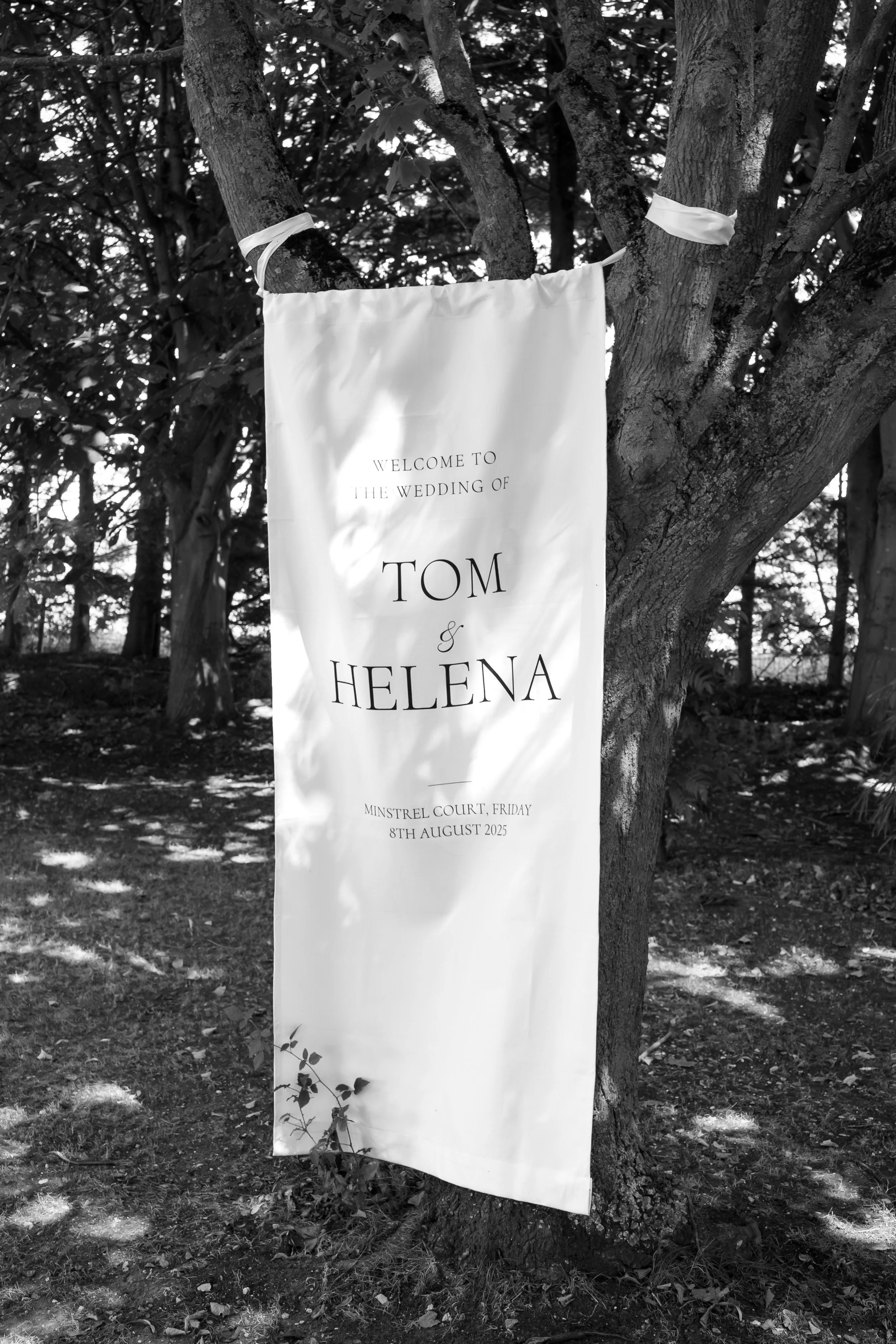 A white banner hanging from a tree with the text 'Welcome to the wedding of Tom & Helena,' indicating a wedding event on August 8th, 2025, at Minstrel Court, in a wooded outdoor setting.