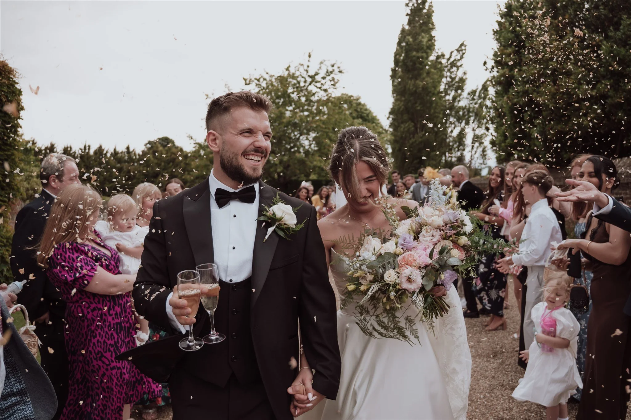 A newlywed couple walking outdoors at their wedding celebration, surrounded by guests, with the groom holding a glass of champagne and the bride holding a large bouquet of flowers, confetti falling around them.