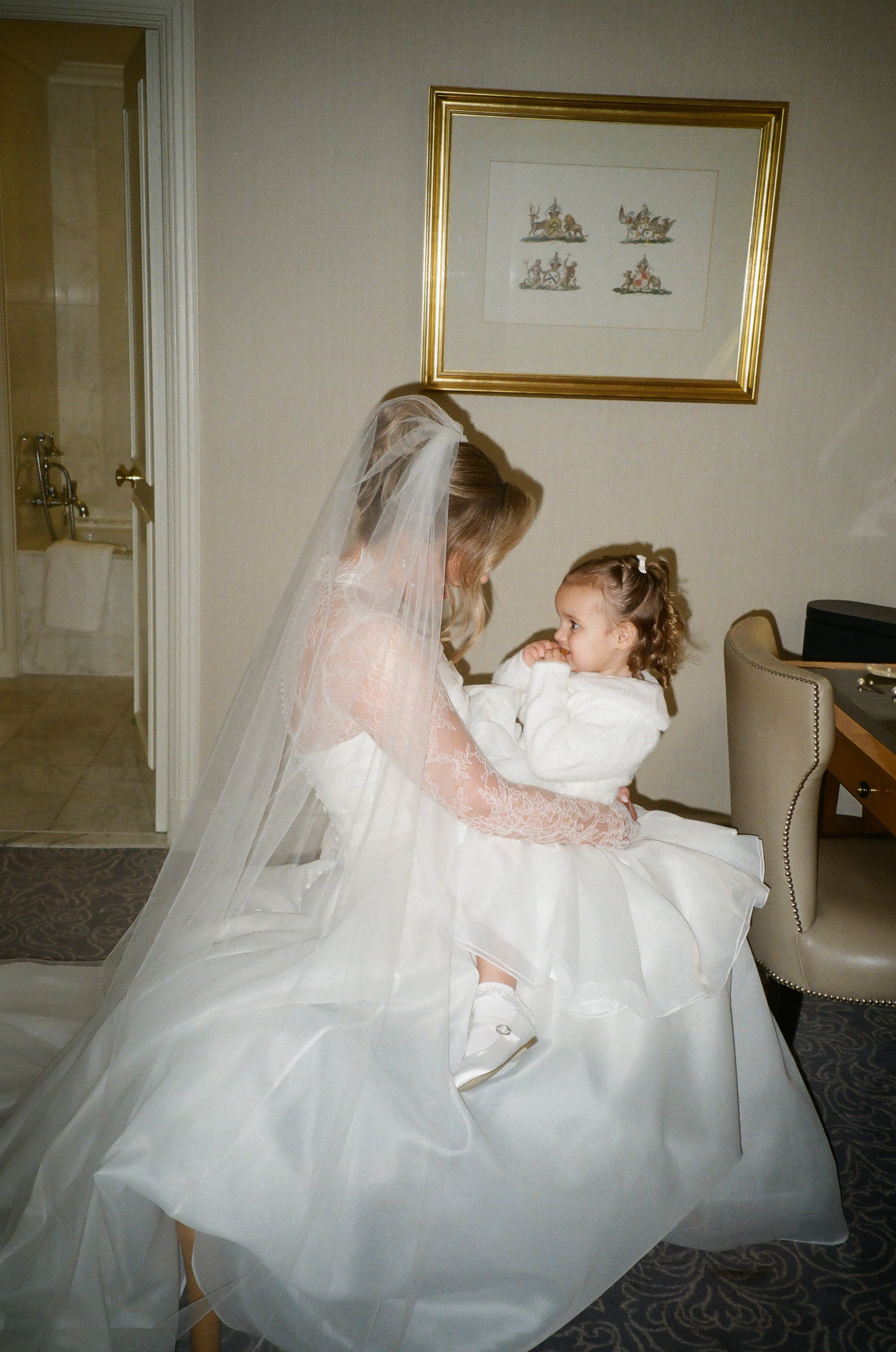 A woman dressed as a bride with a veil sitting on a chair and holding a young girl in a white dress in a hotel room.
