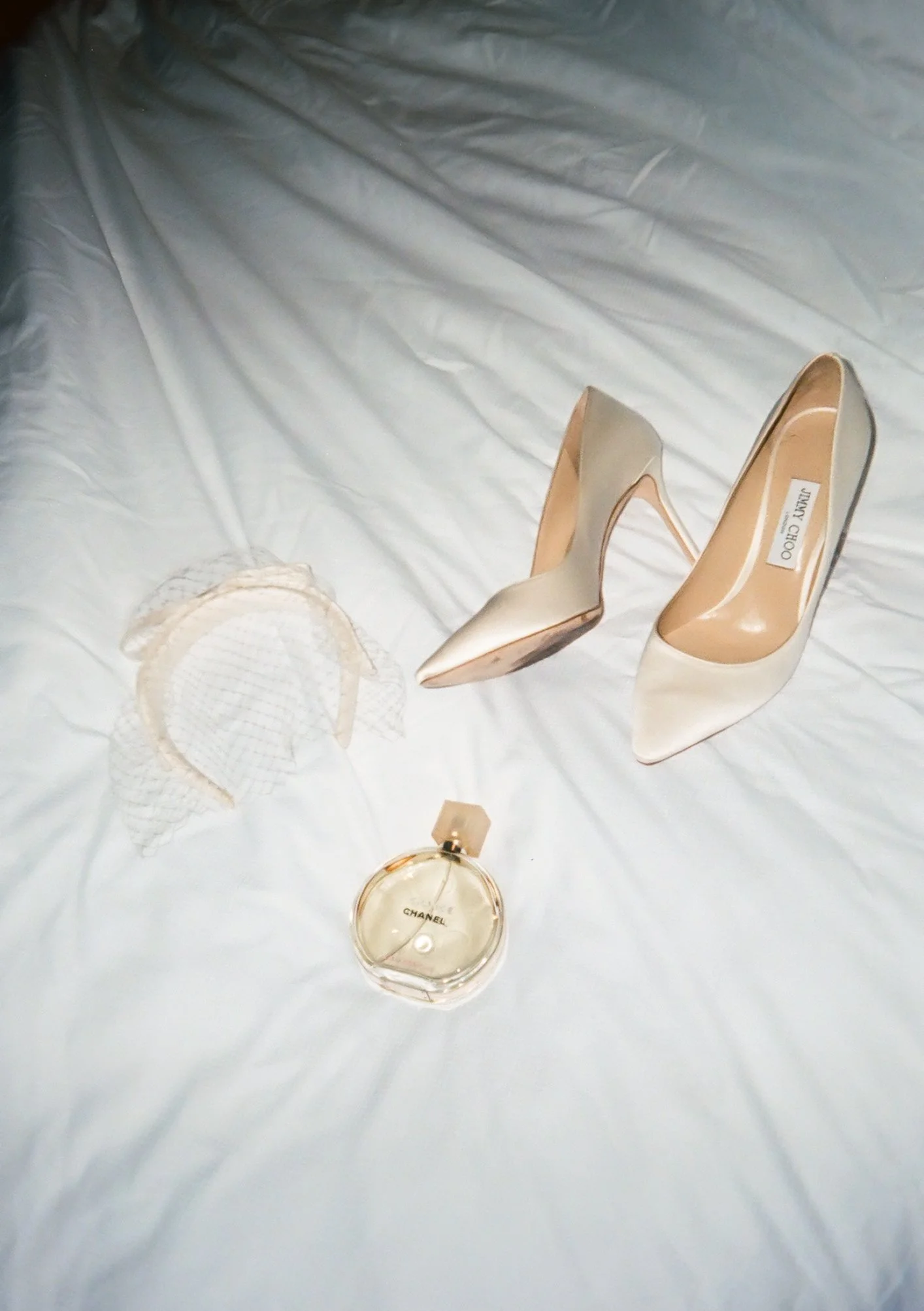 A pair of beige high-heeled shoes, bottle of Chanel perfume, and a small beige clutch purse on a white bedsheet.