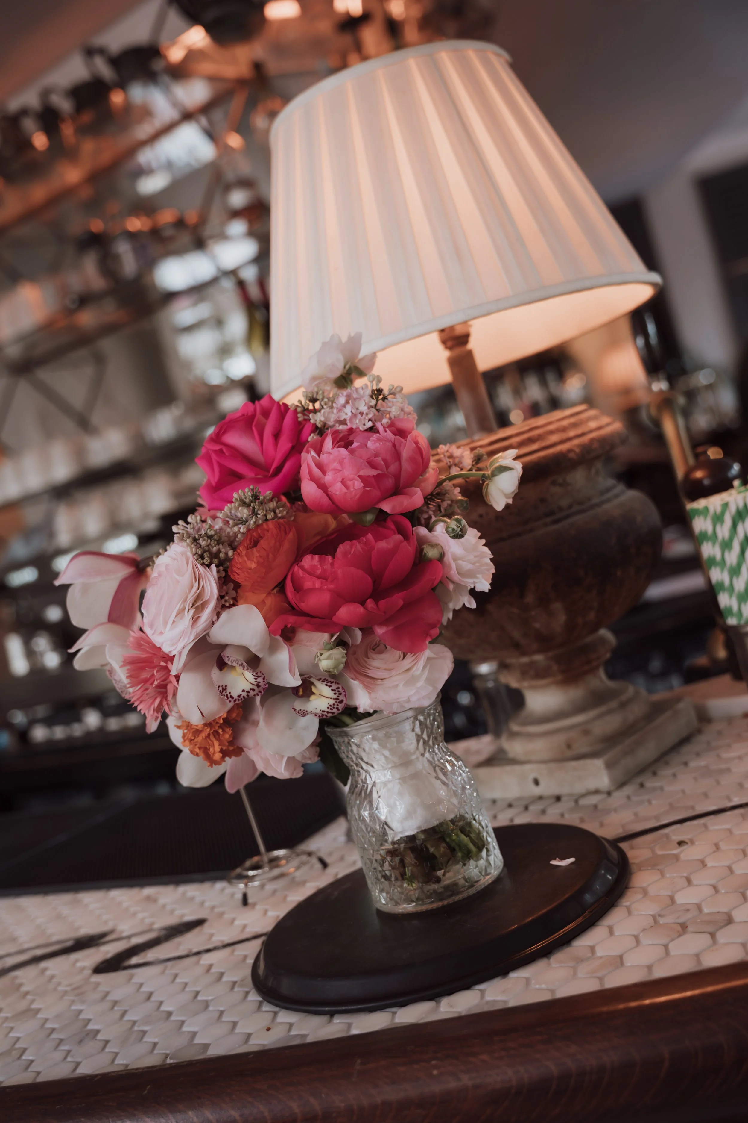 A floral arrangement of pink and white flowers in a glass vase on a wooden tray, with a table lamp with a cream-colored shade and a ribbed design in the background.