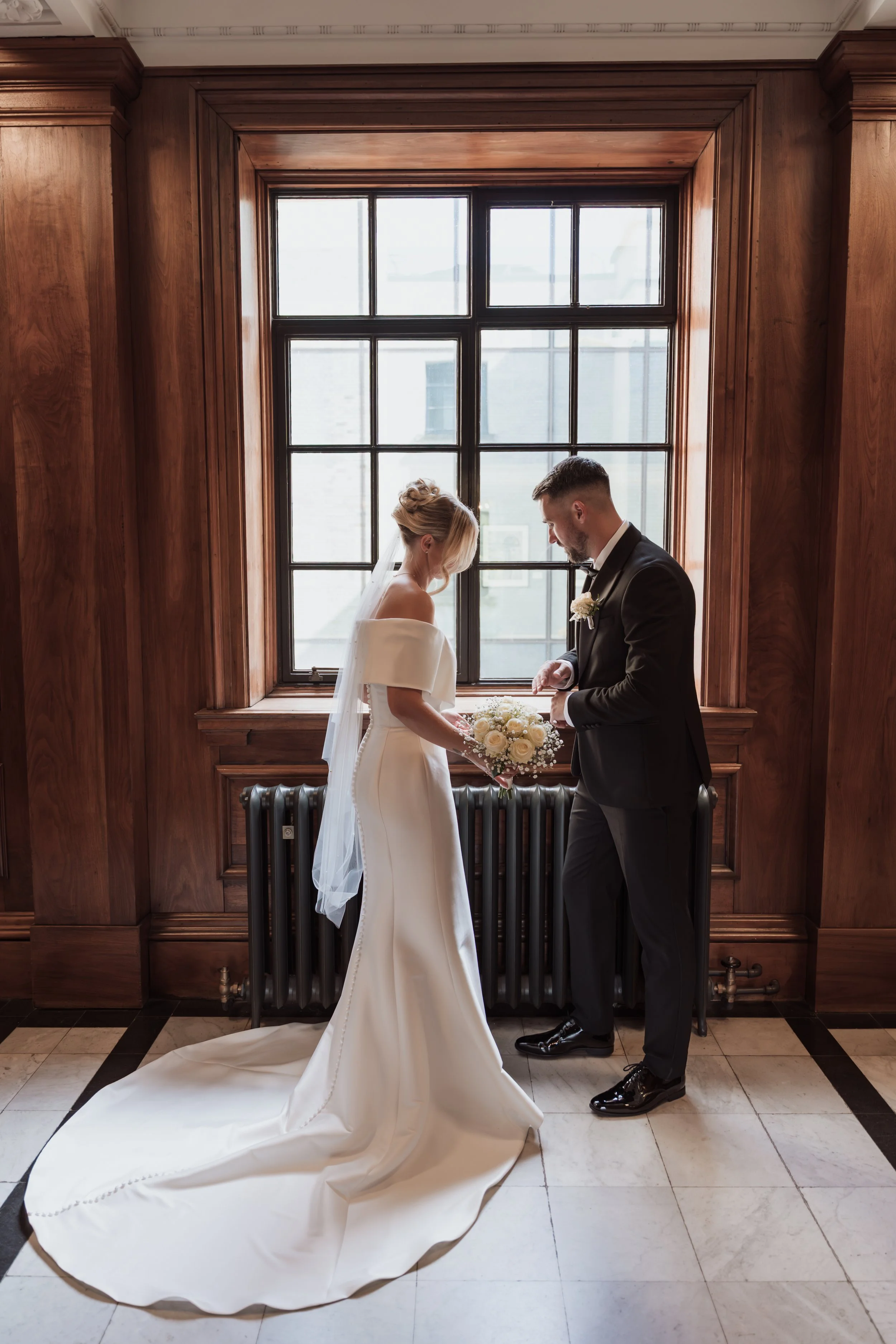 Bride and groom standing in front of a large window with wood frame, exchanging wedding vows. The bride wears a white wedding gown with a train and veil, holding a bouquet of white roses. The groom wears a black tuxedo with a boutonniere, and they ar