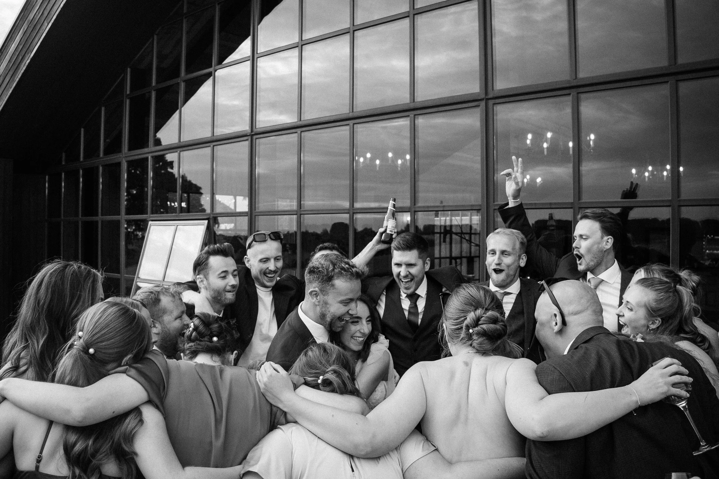 A group of people in formal attire hugging and celebrating outside near a modern building with large glass windows.