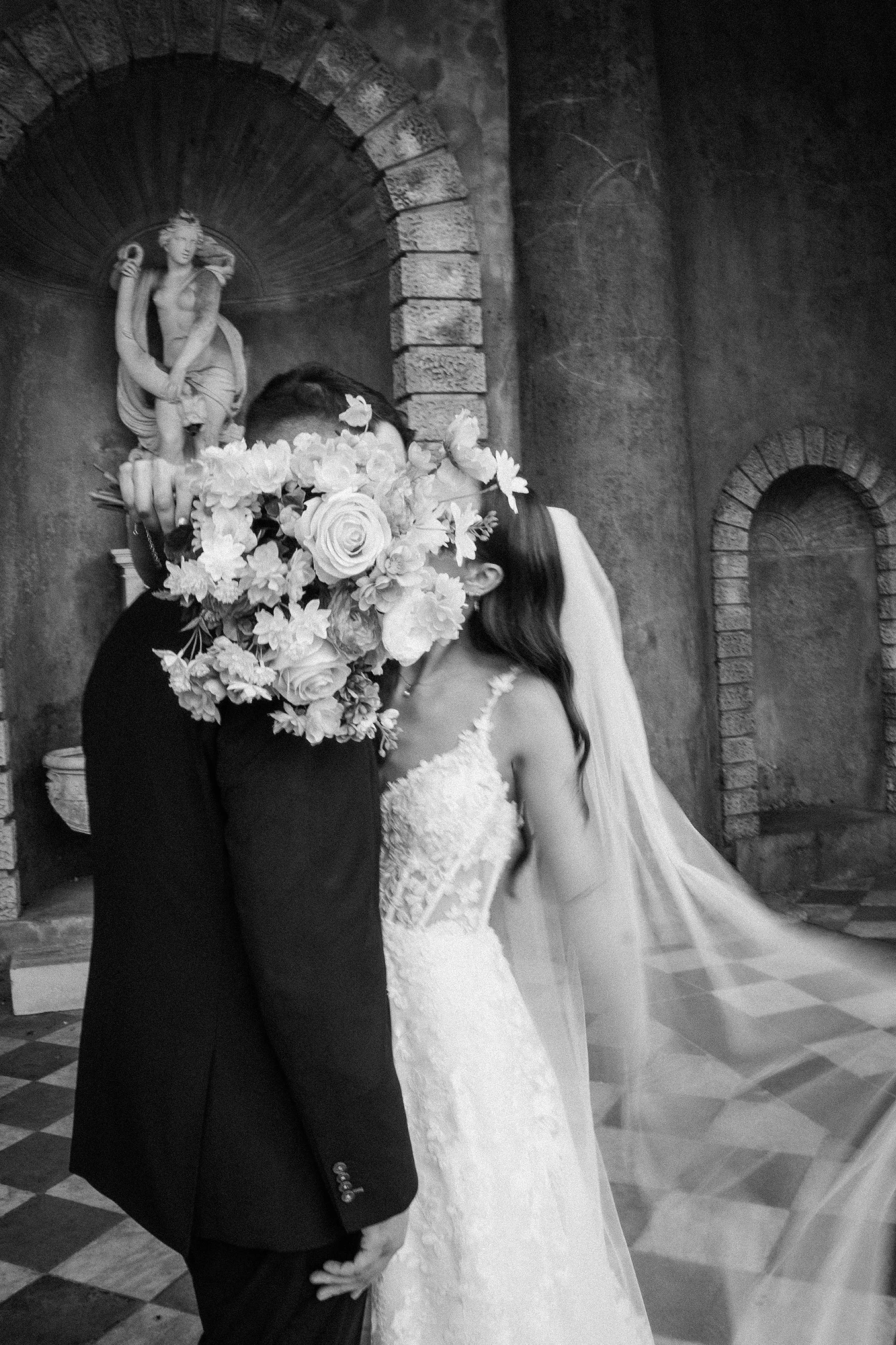 A bride and groom are embracing in a wedding photo, with the groom's face hidden behind a bouquet of flowers held by the bride. The bride is wearing a white dress and the groom is dressed in a dark suit. In the background, there is a small statue of a woman holding a jug, placed in an alcove with brick archways.