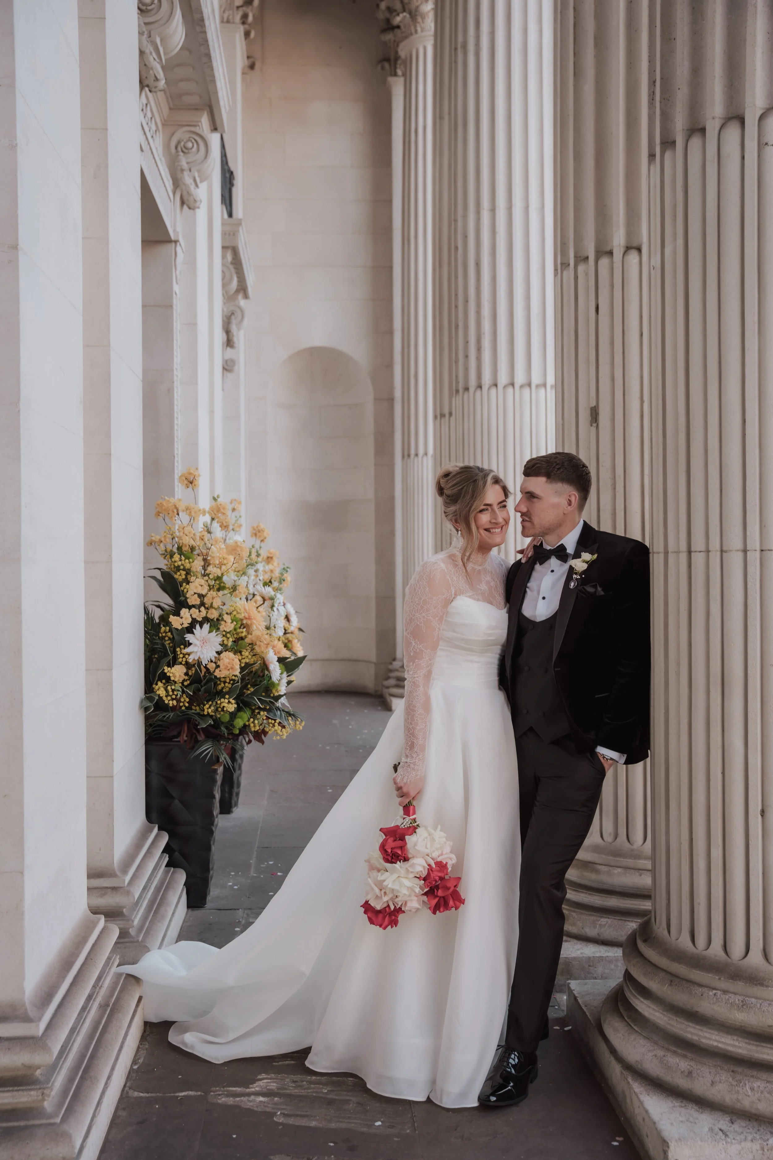 A bride and groom in wedding attire standing close together in front of large stone columns in a historic building. The bride is smiling, holding a bouquet of red and white flowers, and wearing a white gown with lace sleeves. The groom is leaning aga