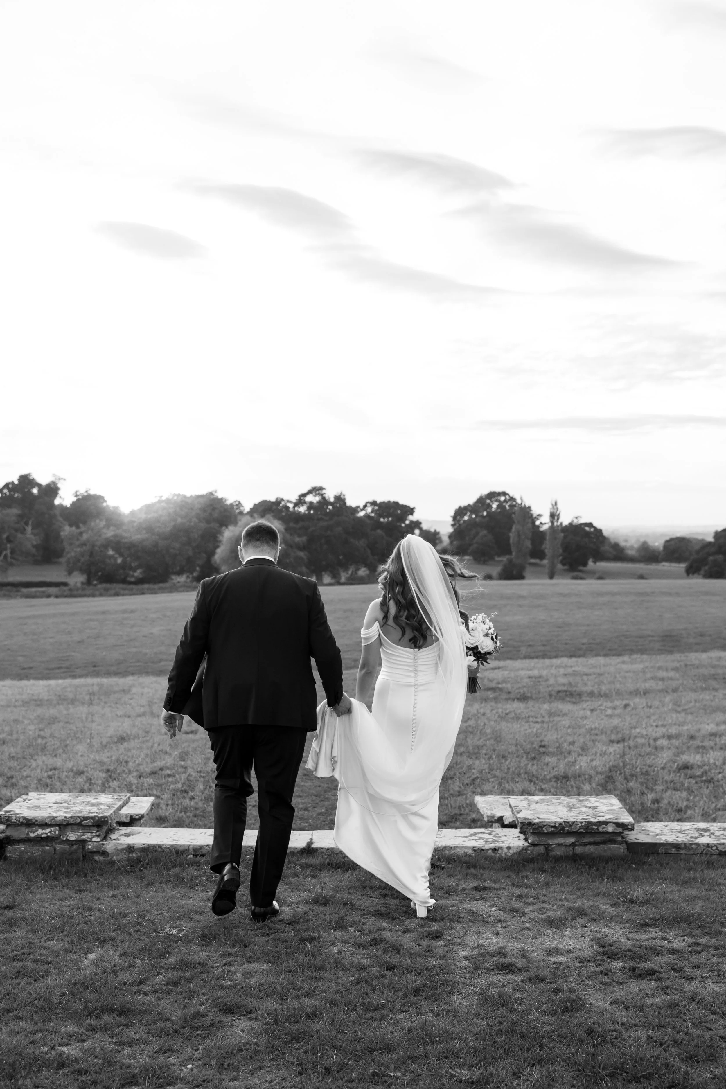 A bride and groom holding hands, walking away outdoors on a grass field during a wedding, with trees and a cloudy sky in the background, in black and white.