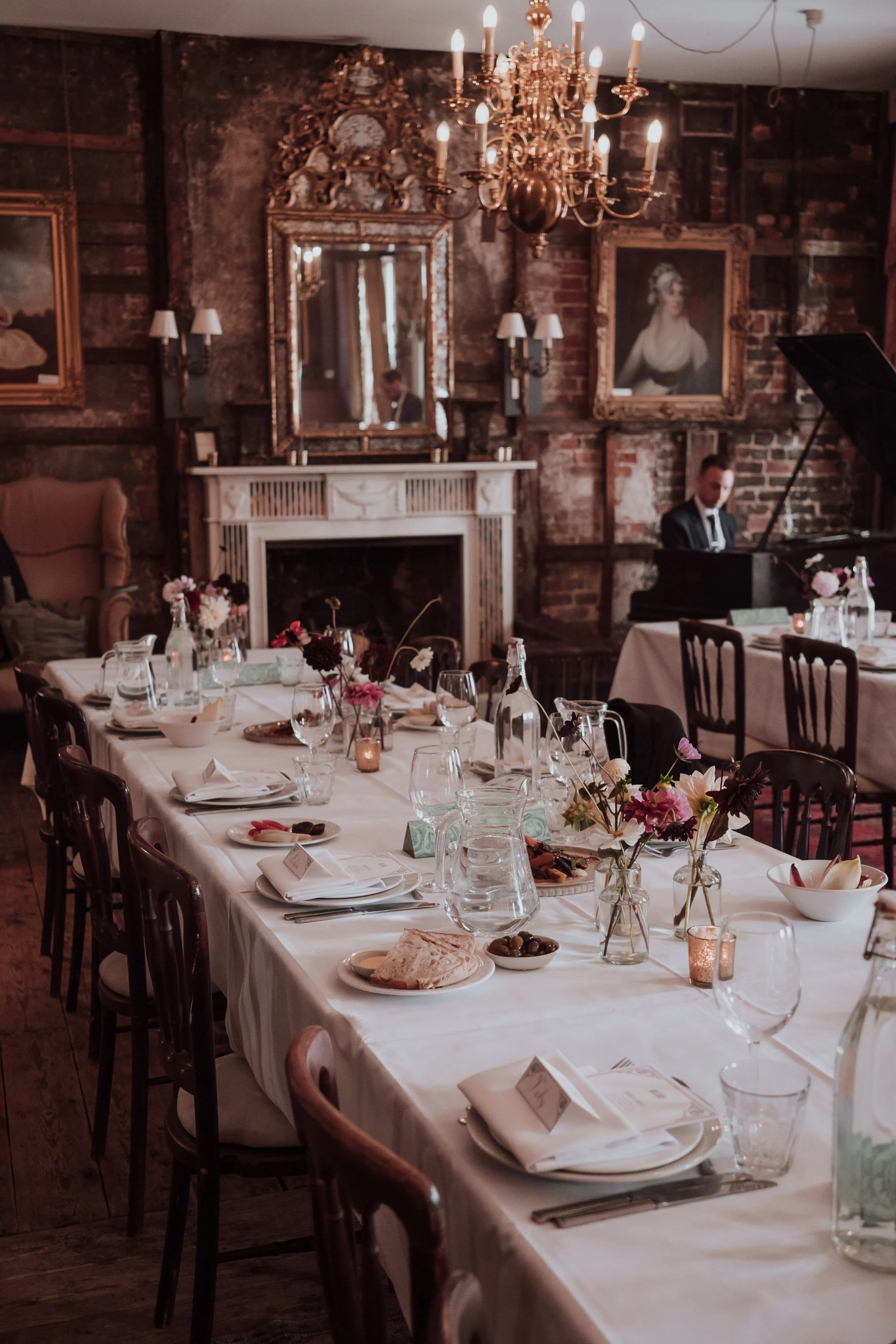 A decorated dining table set for an event with flowers, plates, glasses, and napkins in a vintage-style room with a chandelier, paintings, and a man playing the piano.