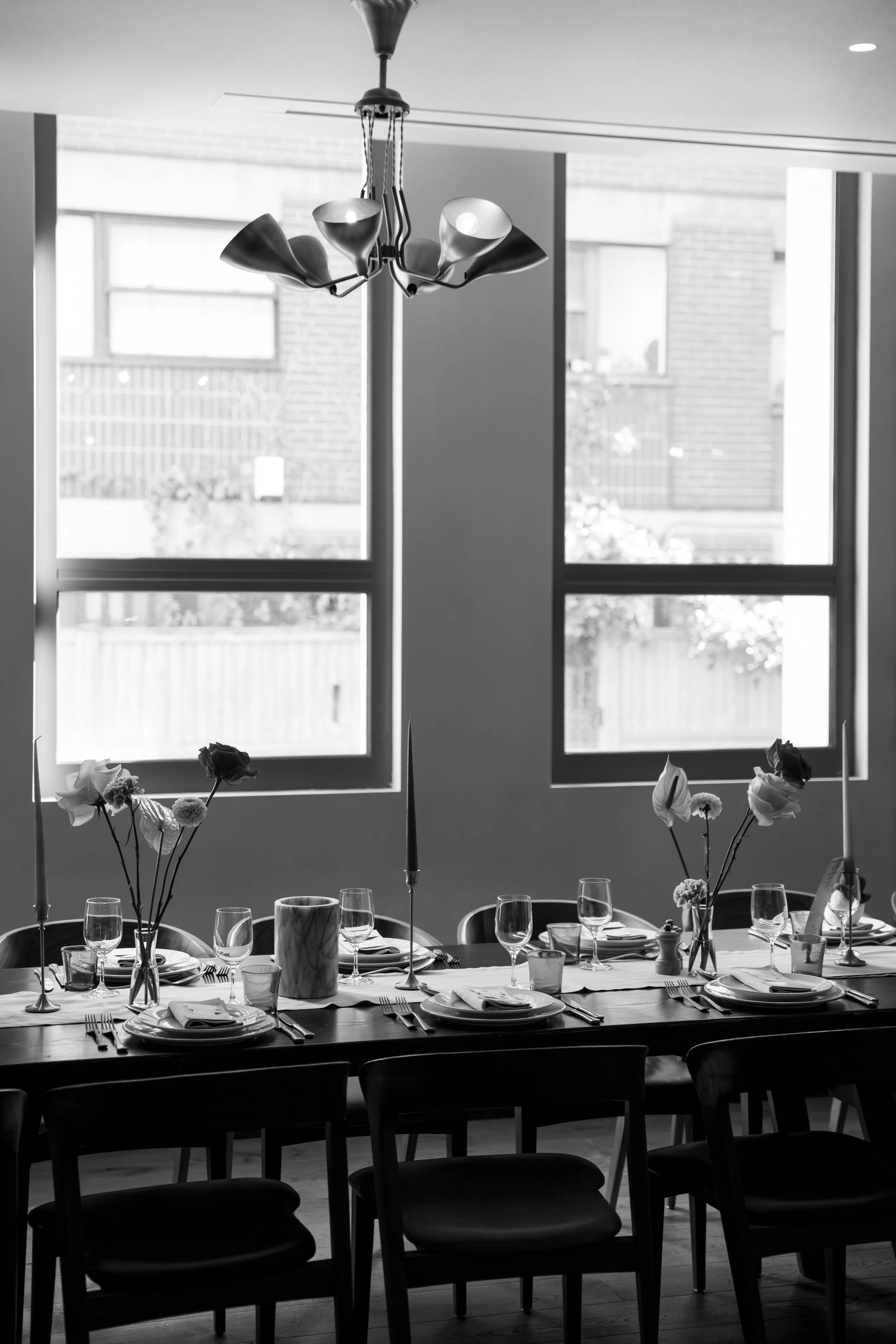 A dining table set for a formal meal with flowers, plates, glasses, and utensils. Large windows with daylight outside and a modern ceiling light fixture.