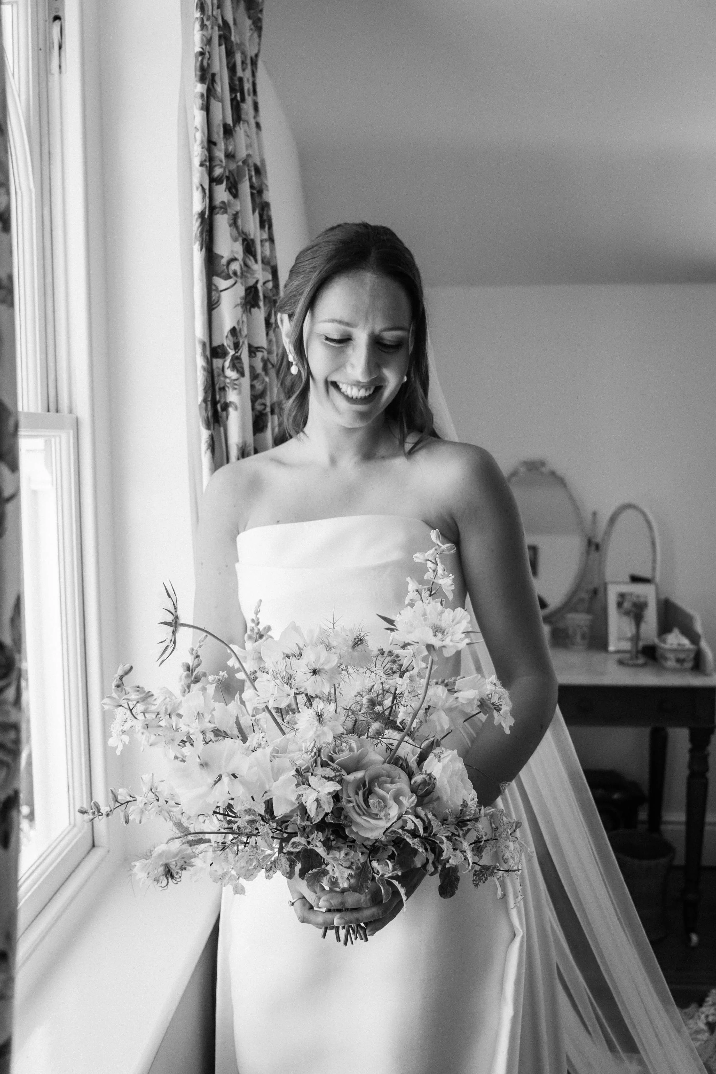 A bride holding a large bouquet of flowers inside a room, smiling with her eyes closed.