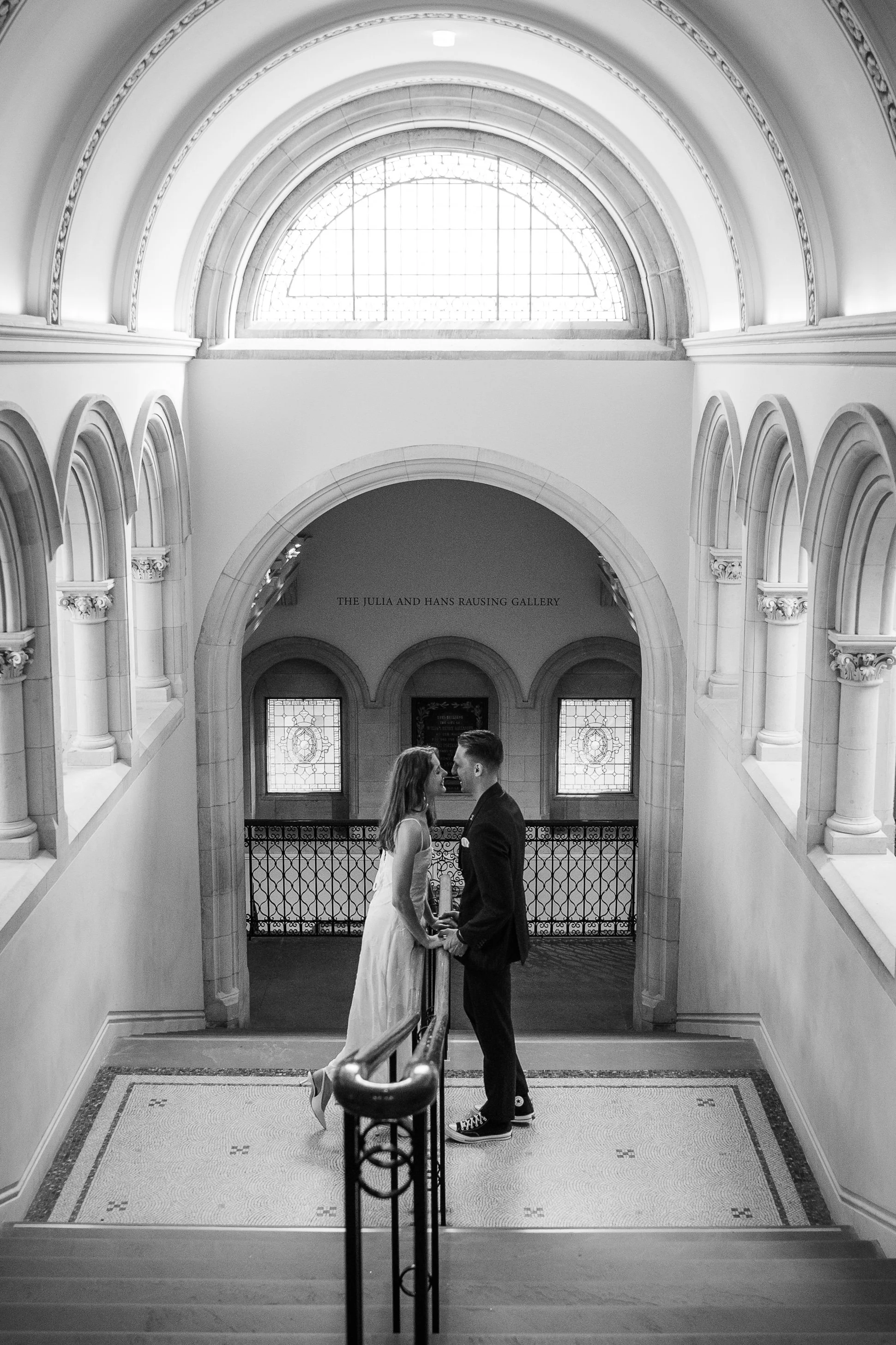 A black and white photo of a bride and groom standing in a historic building, holding hands and facing each other, underneath an arched window with natural light streaming through. The architecture features ornate columns and detailed molding.