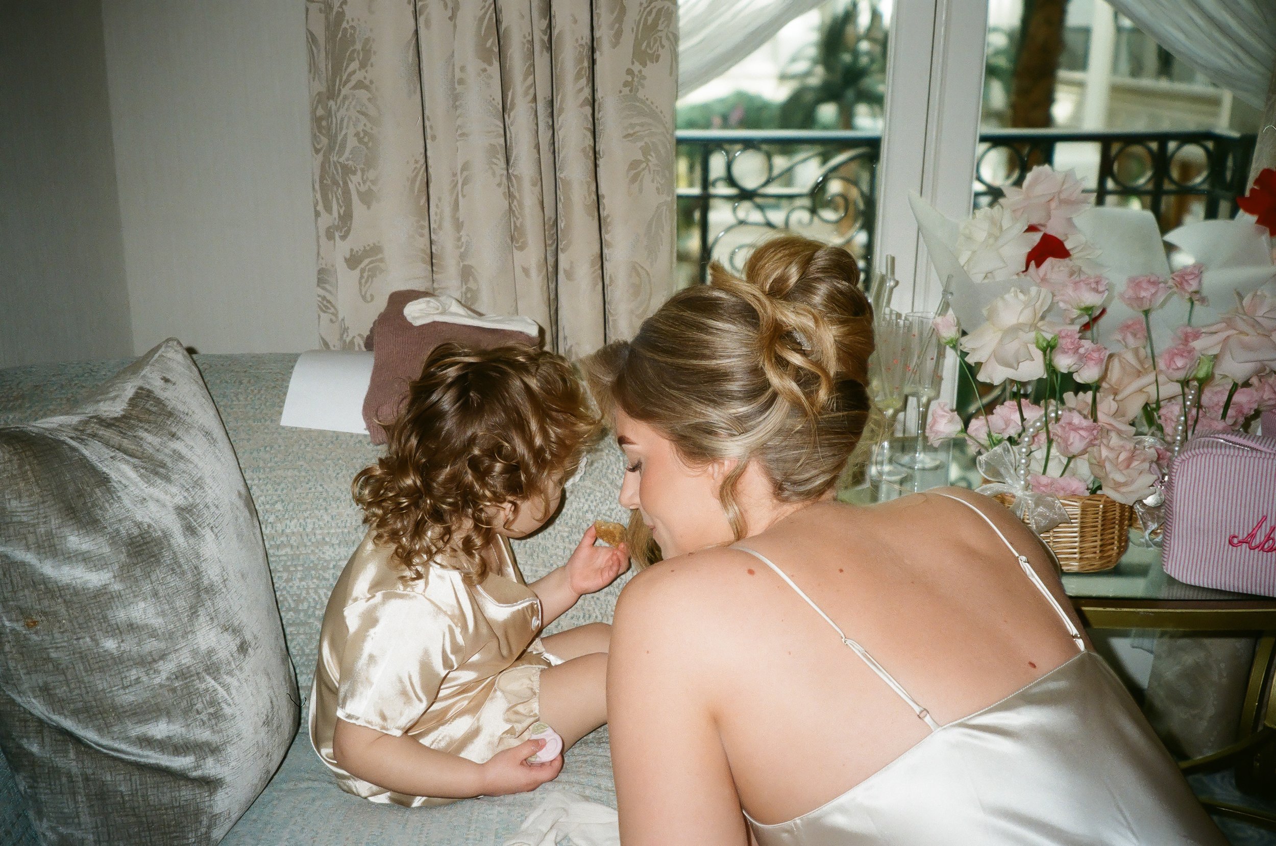 A young girl and a woman are sitting on a sofa, leaning towards each other, sharing a moment. The girl is wearing a satin dress and has curly hair, while the woman, possibly her mother, is in a sleepwear top with spaghetti straps. Behind them, there are decorated tables with pink and white flowers, a basket, and glasses on display, suggesting a celebration or special occasion. A window with floral curtains and a balcony railing are visible in the background.