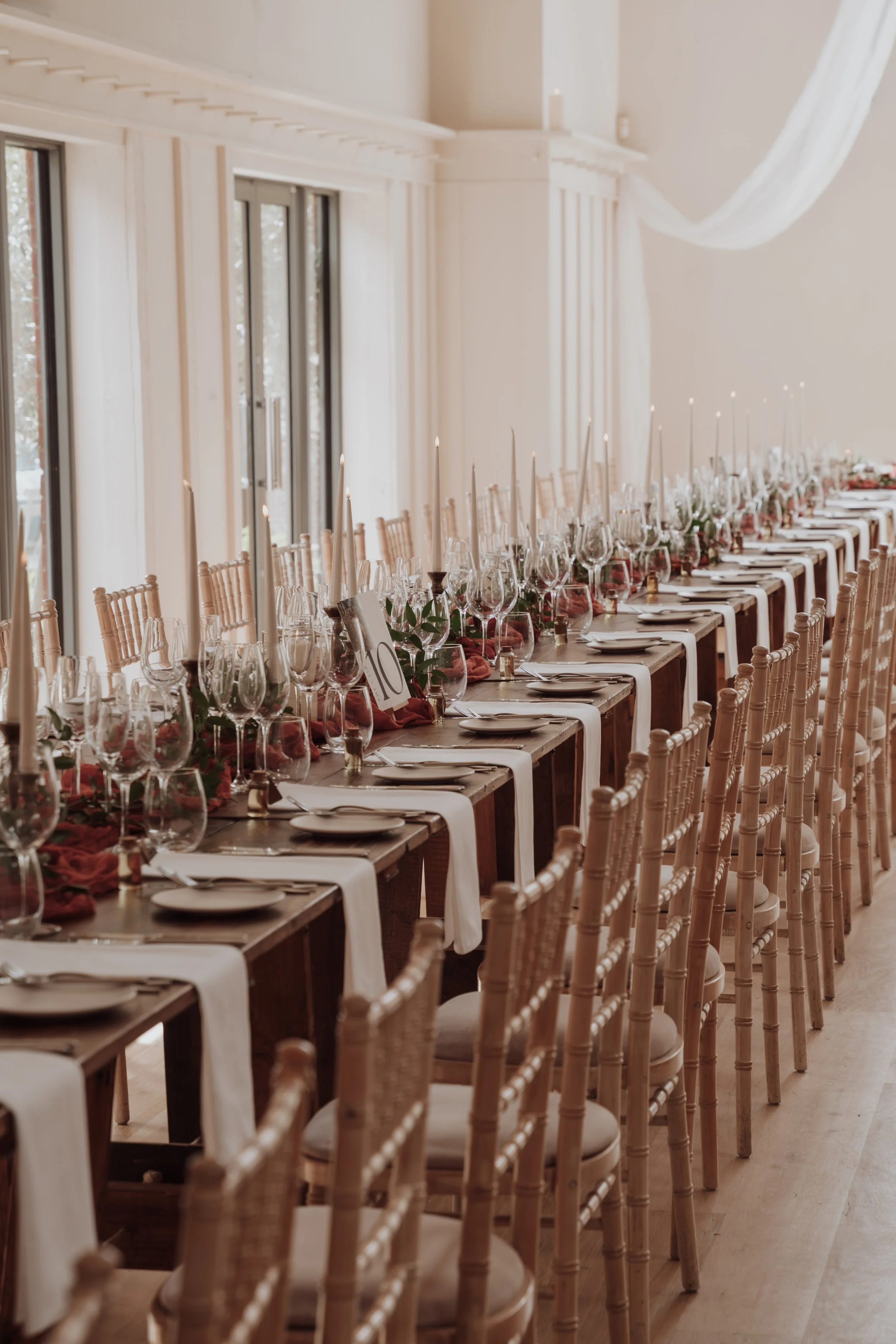 Long banquet table set with wine glasses, candles, flowers, and place settings in a bright, elegant room with large windows and white walls.