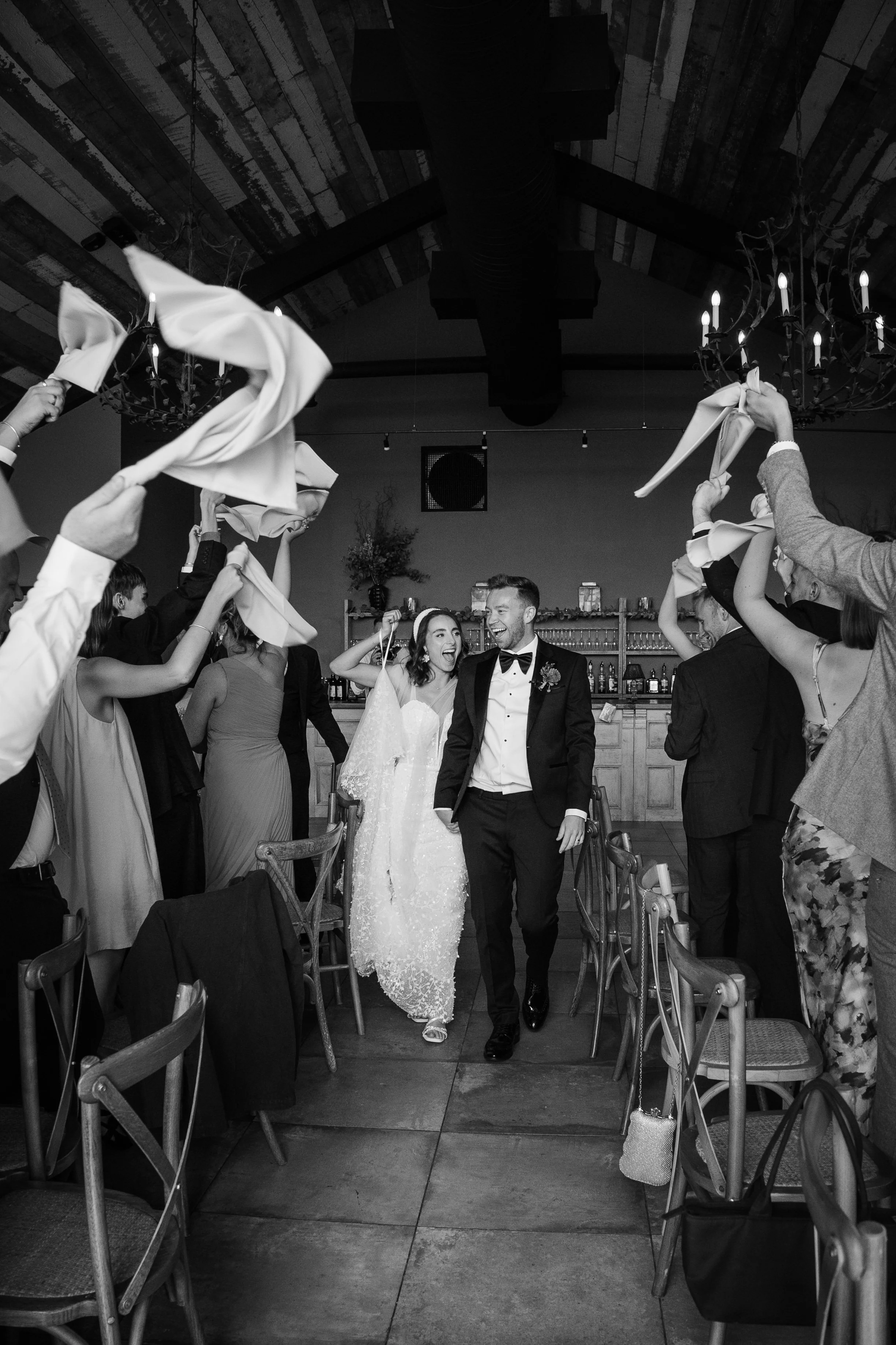 A black and white photo of a wedding celebration with a bride and groom walking through a dance floor surrounded by guests holding napkins and cheering. The setting appears to be an indoor venue with chandeliers and a bar area in the background.