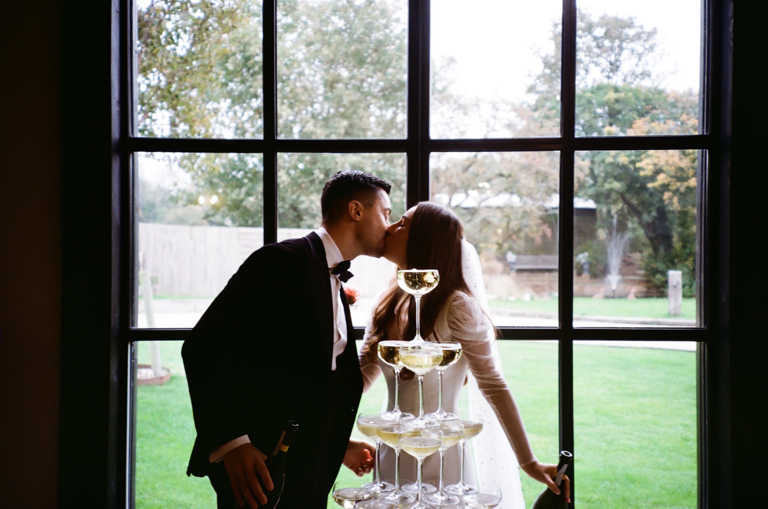 A couple sharing a kiss through a large window during daytime, with stacked champagne glasses on a table in front of them, outside a grassy yard with trees.