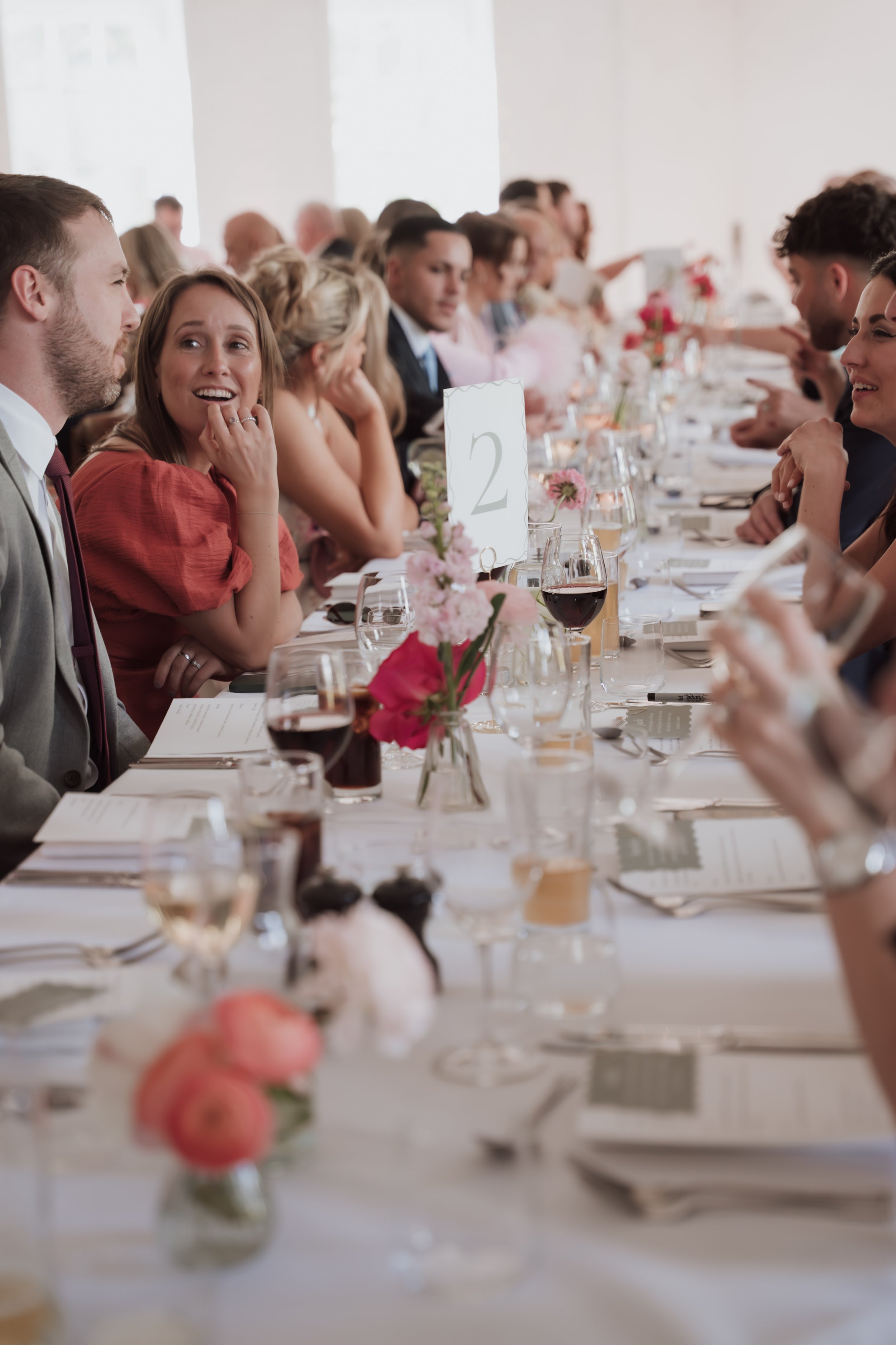People sitting at a long banquet table, engaged in conversation during a formal event or celebration, with floral centerpieces and glasses of wine.