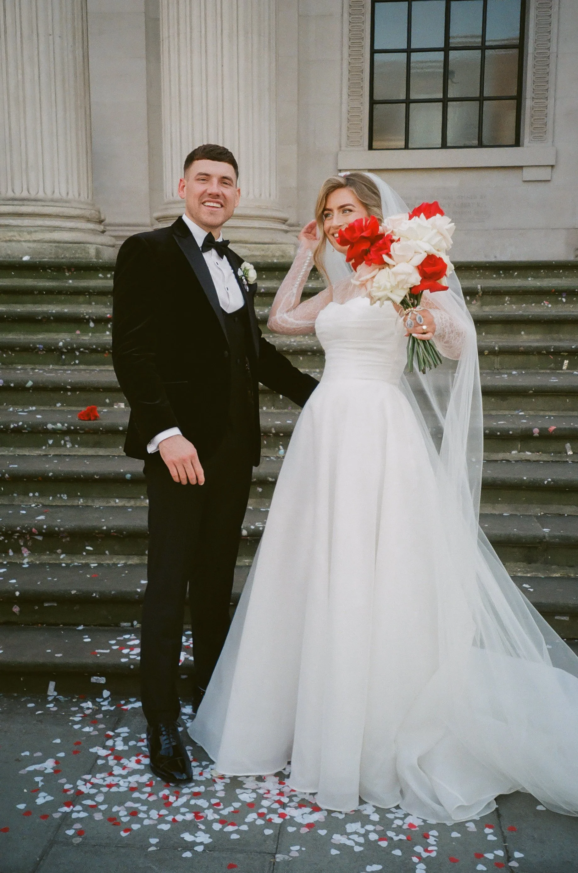 A bride and groom standing on the steps of a church, celebrating their wedding. The bride is holding a bouquet of red and white flowers, and they are surrounded by scattered flower petals.
