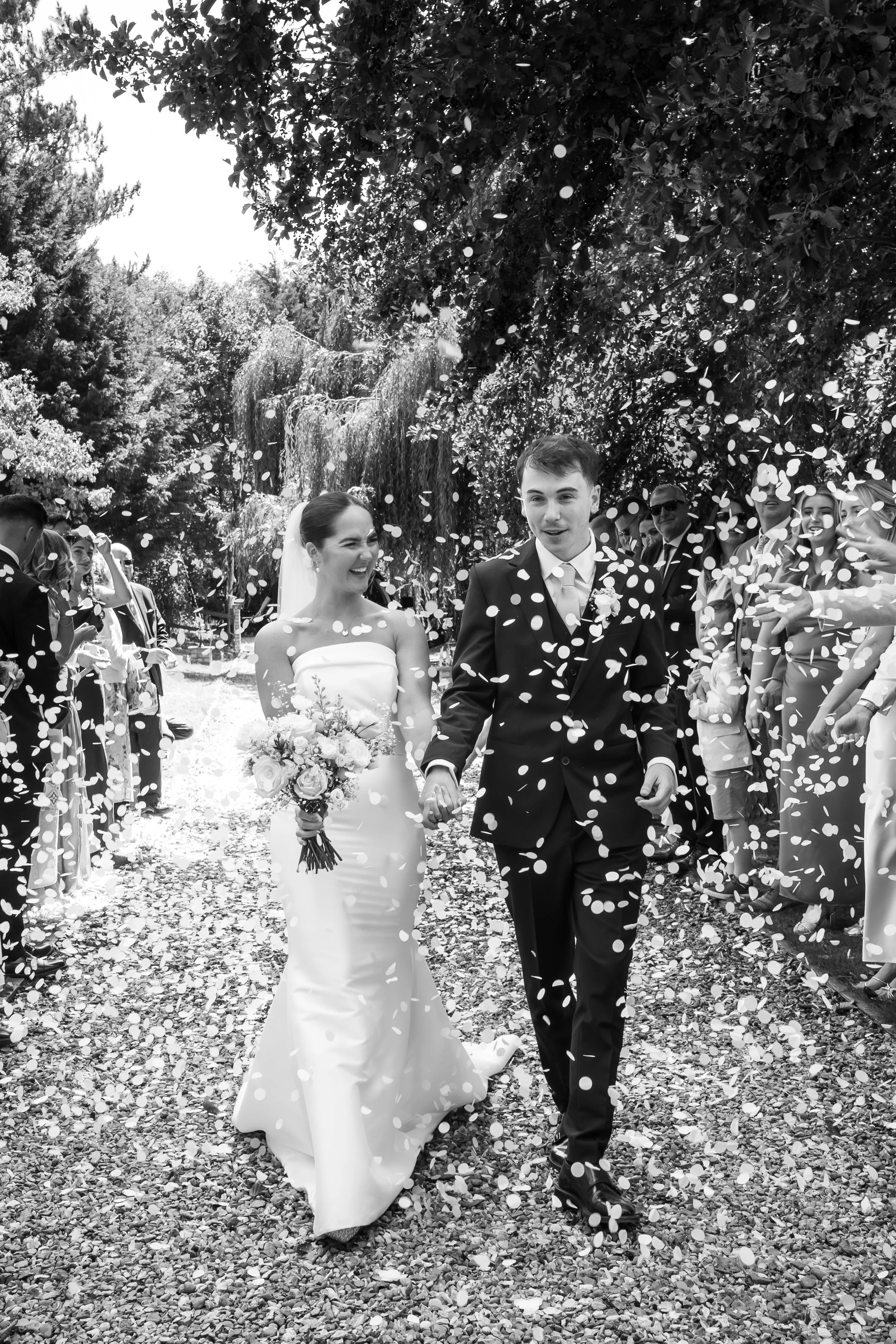 A bride and groom walking down an outdoor path surrounded by guests, with confetti falling around them in a wedding celebration.