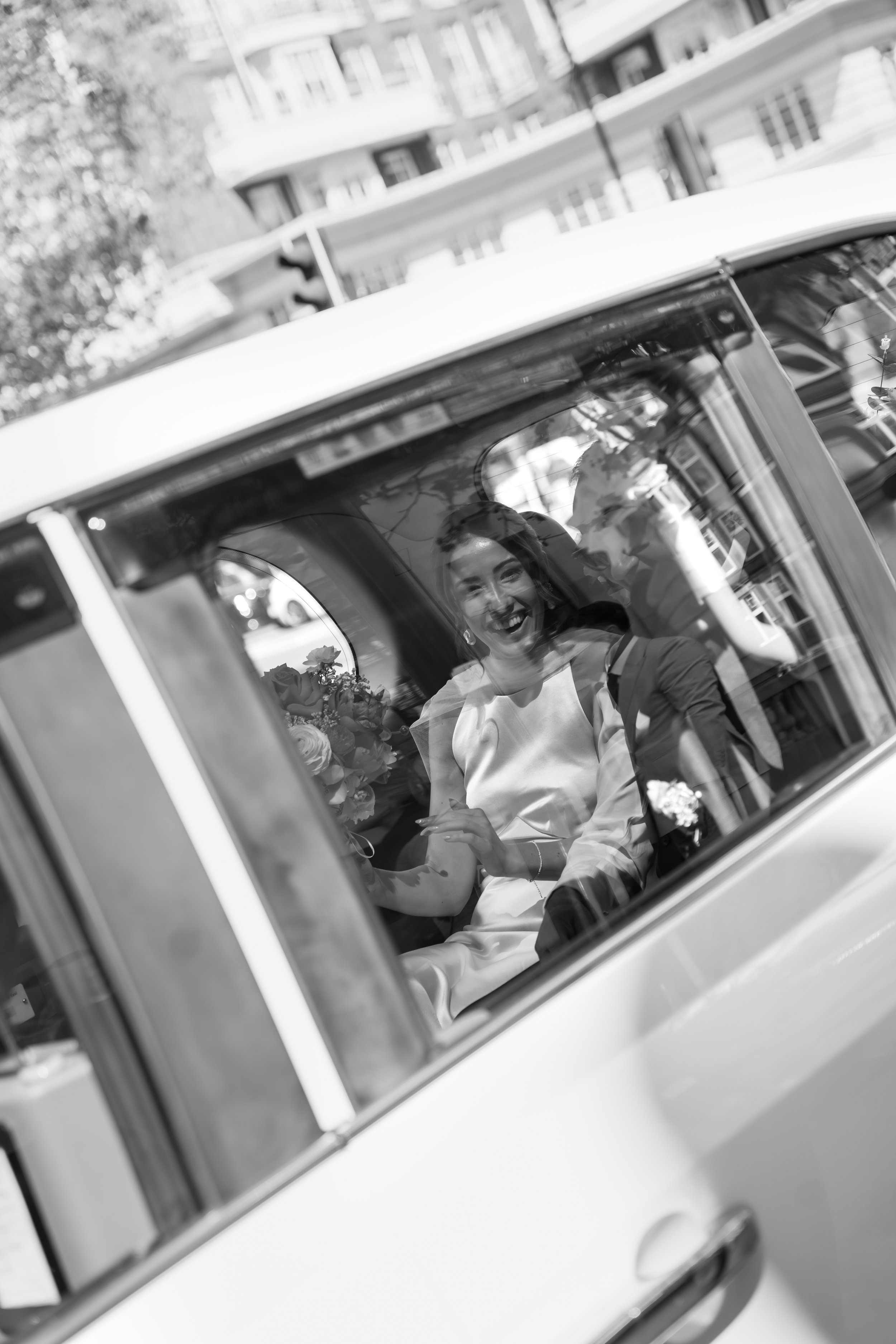 A woman dressed in white, likely a bride, smiling and looking through the window of a vehicle, with a bouquet of flowers visible inside.