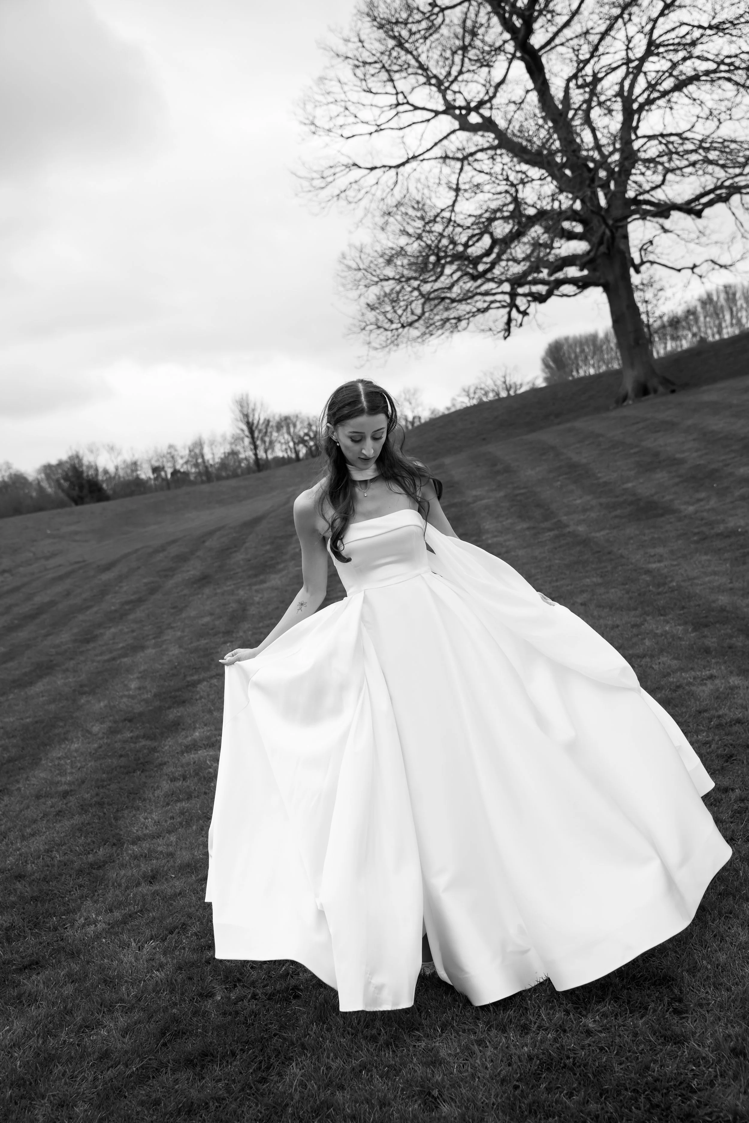 A woman in a white gown standing outdoors on a grassy field, with leafless trees in the background, under an overcast sky.