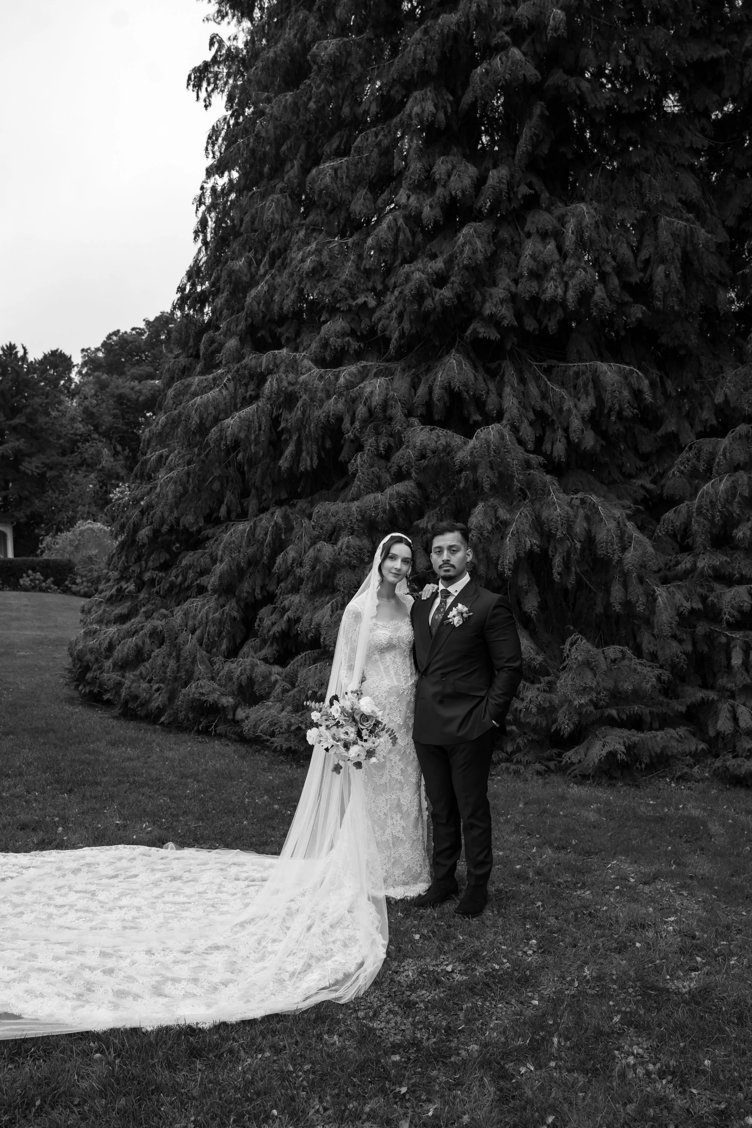 Black and white photo of a bride and groom standing outdoors in front of a large evergreen tree, dressed in wedding attire, with the bride holding a bouquet of flowers.
