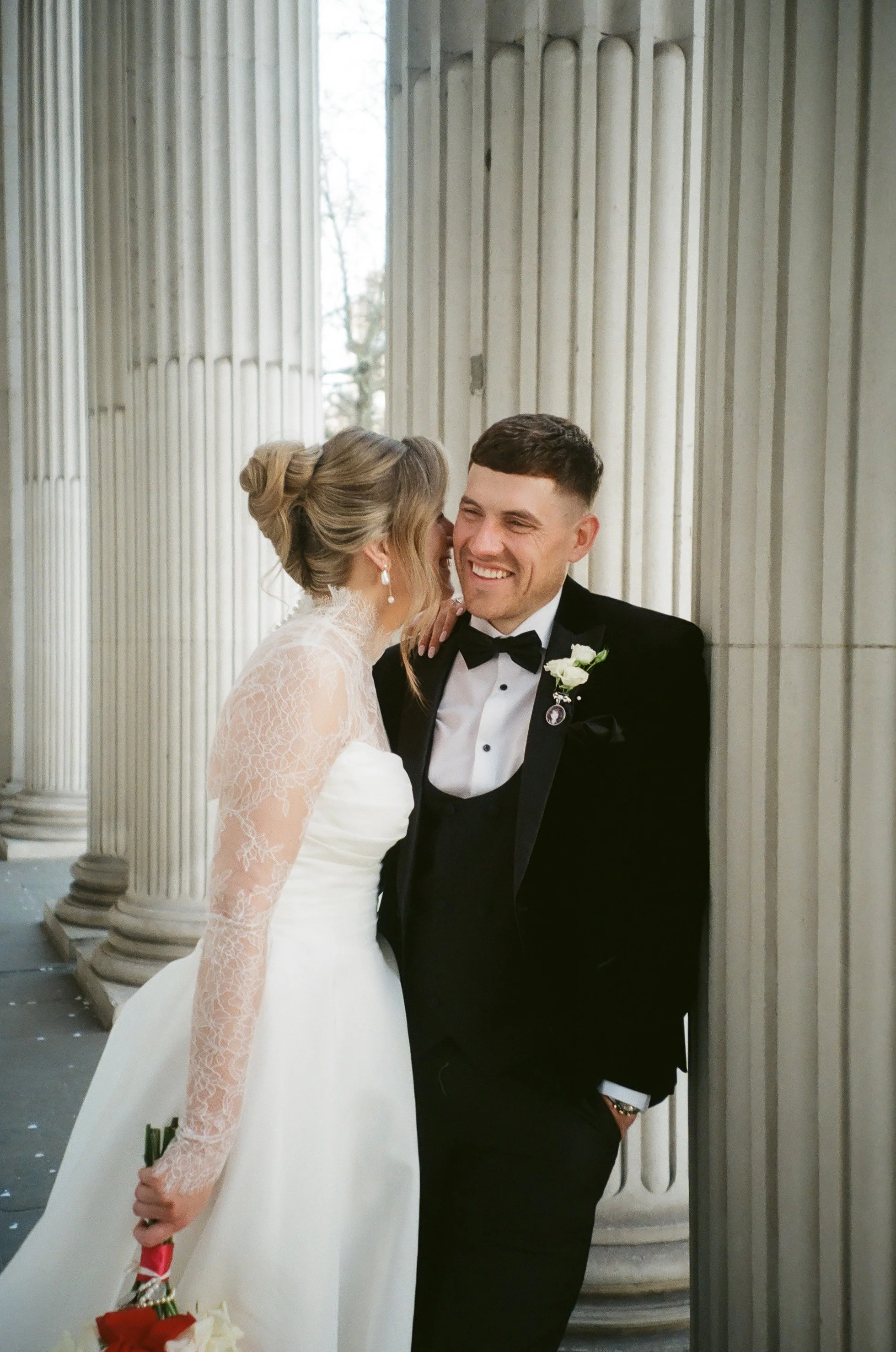A bride and groom standing close together in front of large stone columns, smiling and dressed in wedding attire. The bride wears a white lace dress with long sleeves, and the groom wears a black tuxedo with a bow tie. The bride is holding a bouquet of flowers.