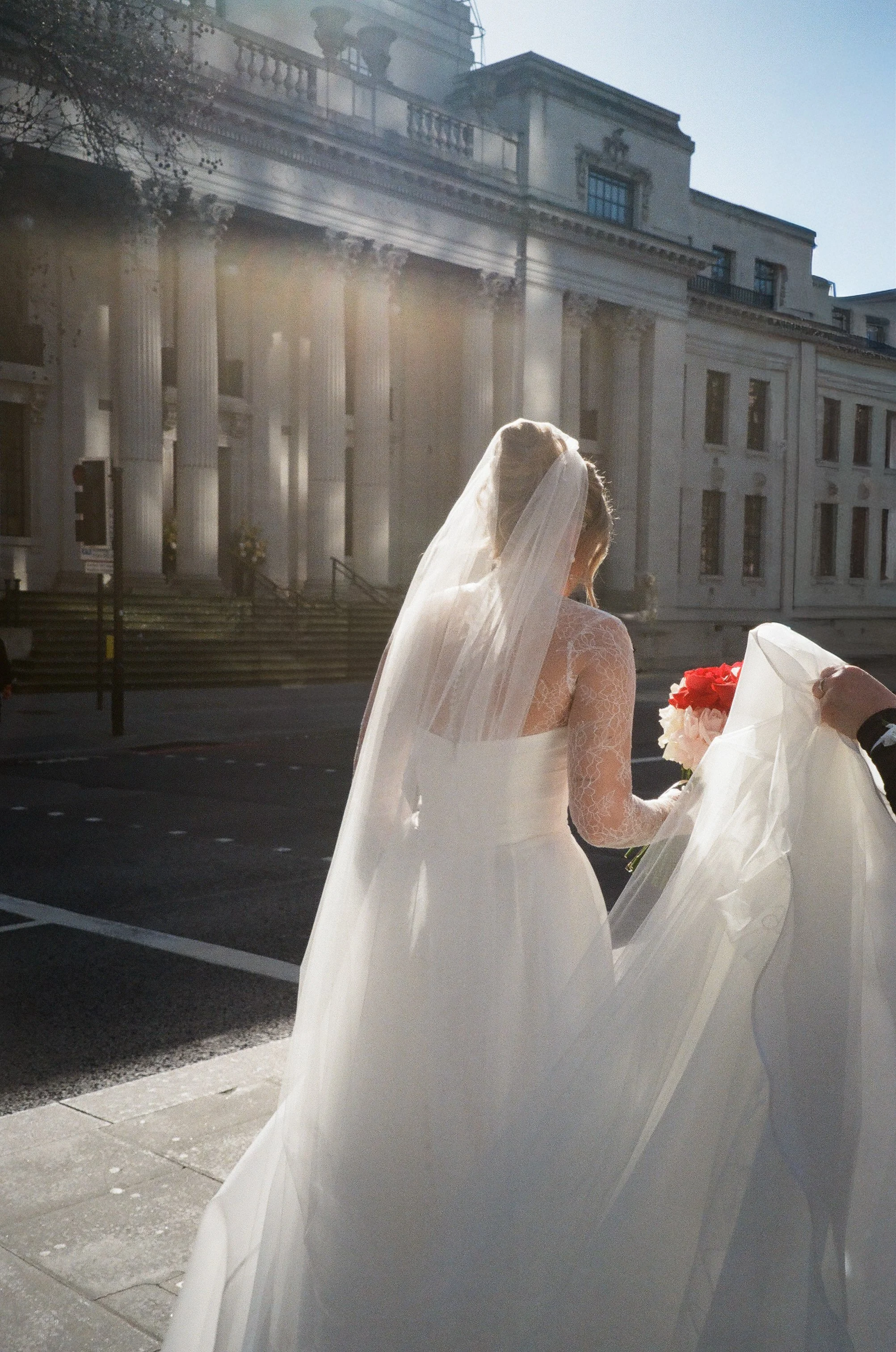 A bride in a wedding dress and veil holding a bouquet of flowers, standing outdoors in front of a grand building with tall columns, with sunlight shining through.