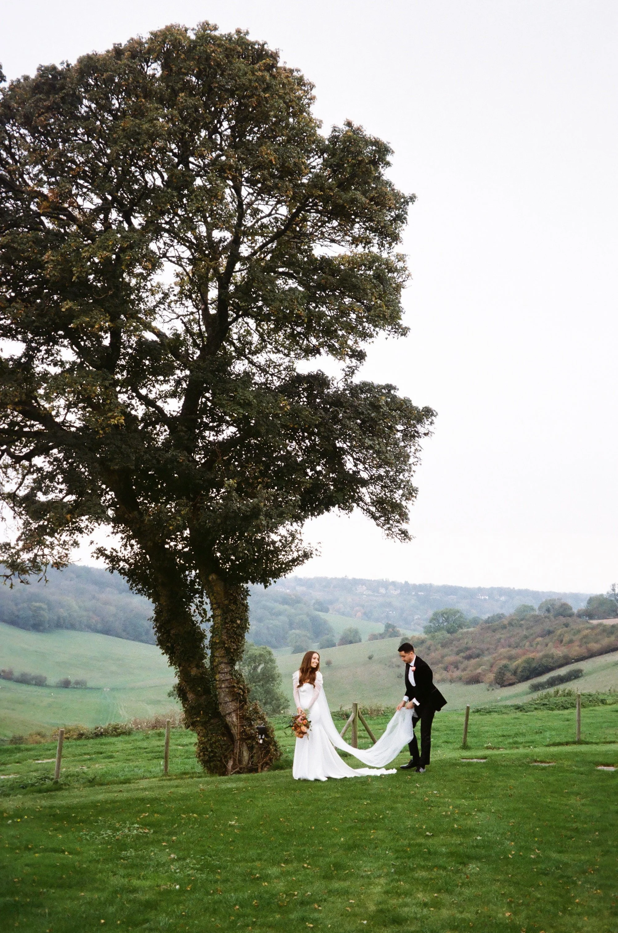 A bride and groom in wedding attire holding hands and smiling under a large tree in a scenic outdoor setting with rolling hills in the background.