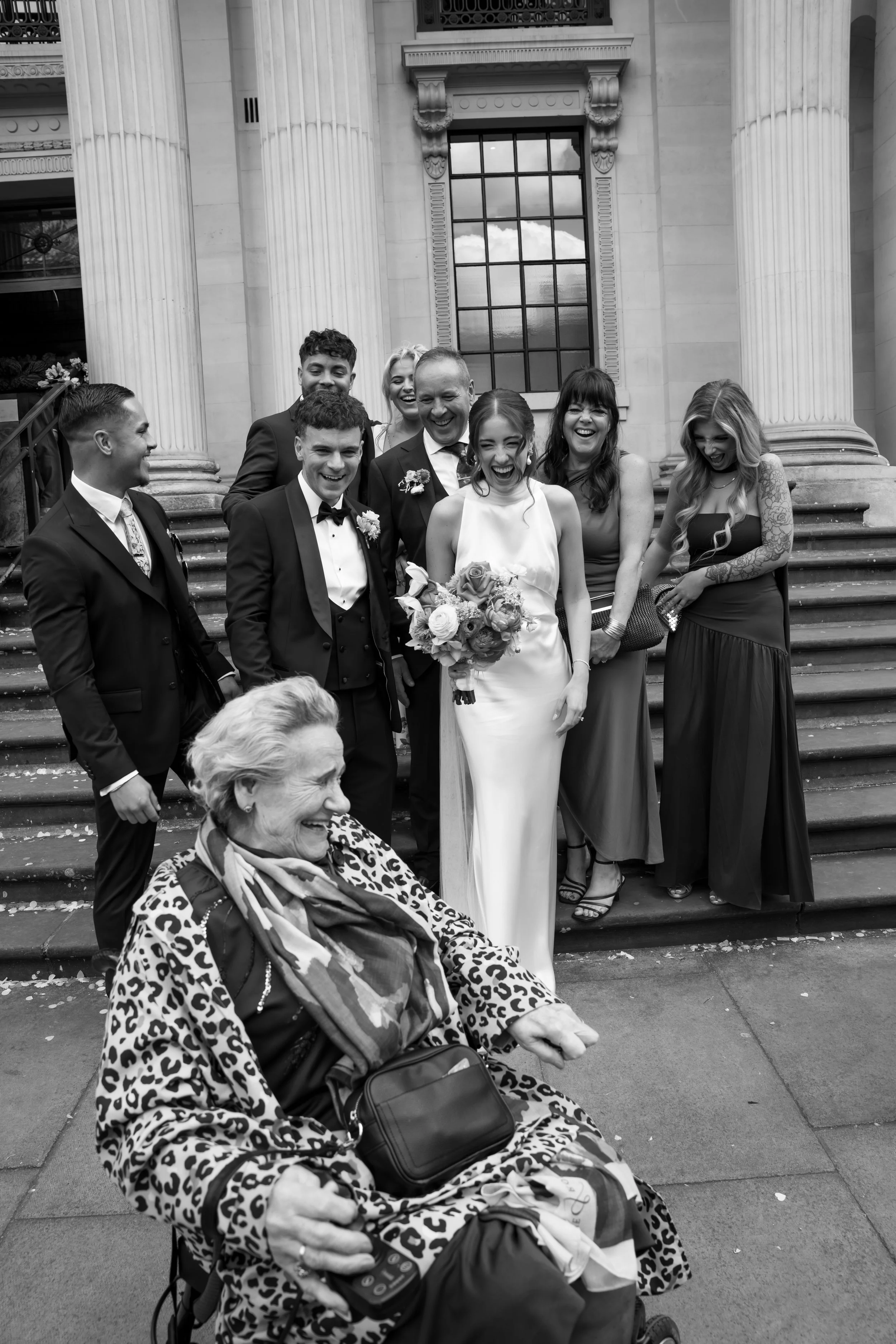 A group of people, including a bride in a wedding dress and several others in formal attire, standing on the steps of a building with large columns and a window reflecting the sky. An elderly woman with a leopard print jacket and a bag is sitting in 