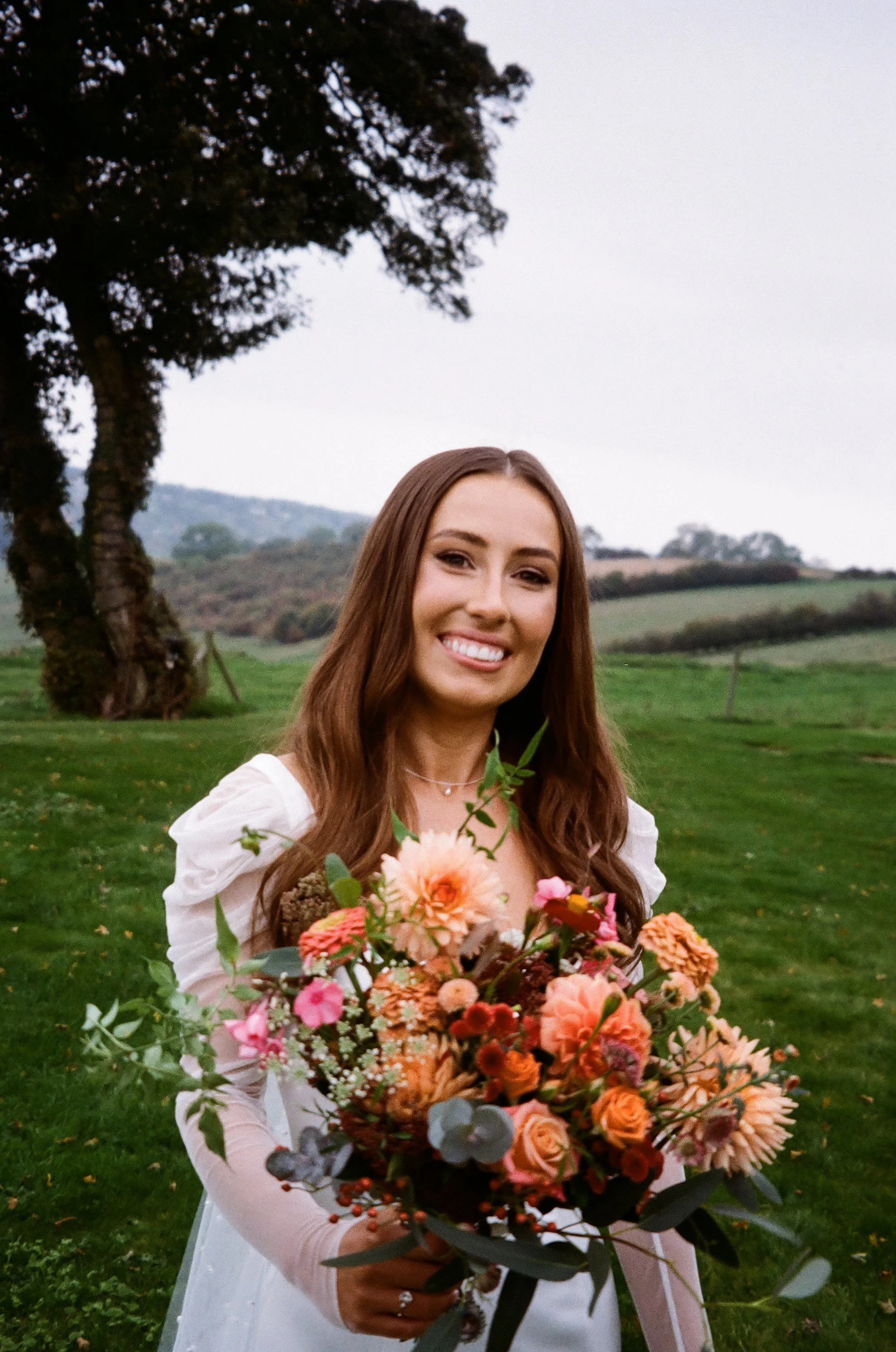 A woman in a white dress holding a large bouquet of colorful flowers outdoors in a green field with trees and hills in the background.