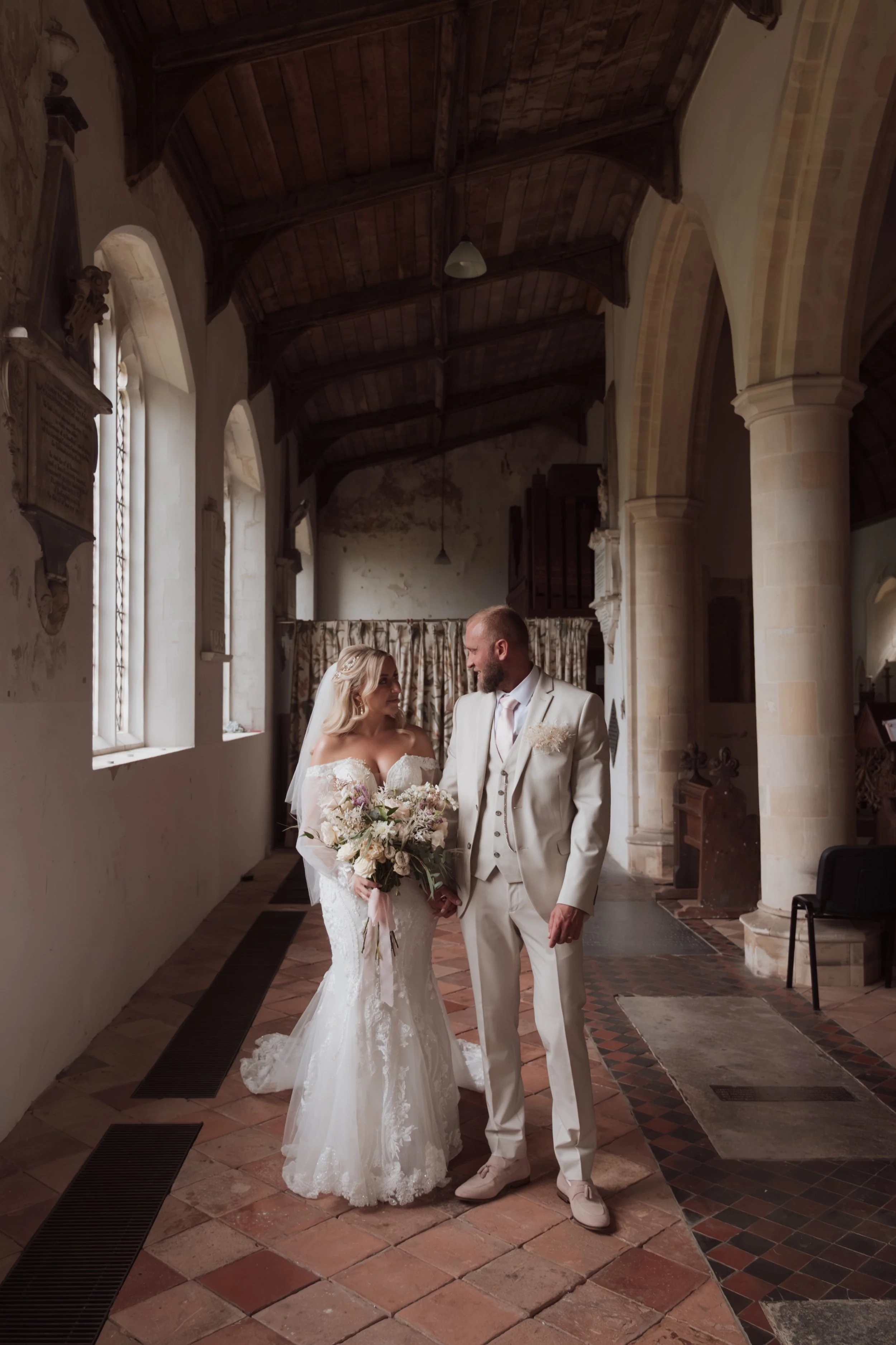 A bride and groom standing in a church with Gothic architecture, wooden ceiling, stone columns, stained glass windows, and vintage decor. The bride wears a lace wedding gown and holds a bouquet, and the groom wears a light-colored suit.