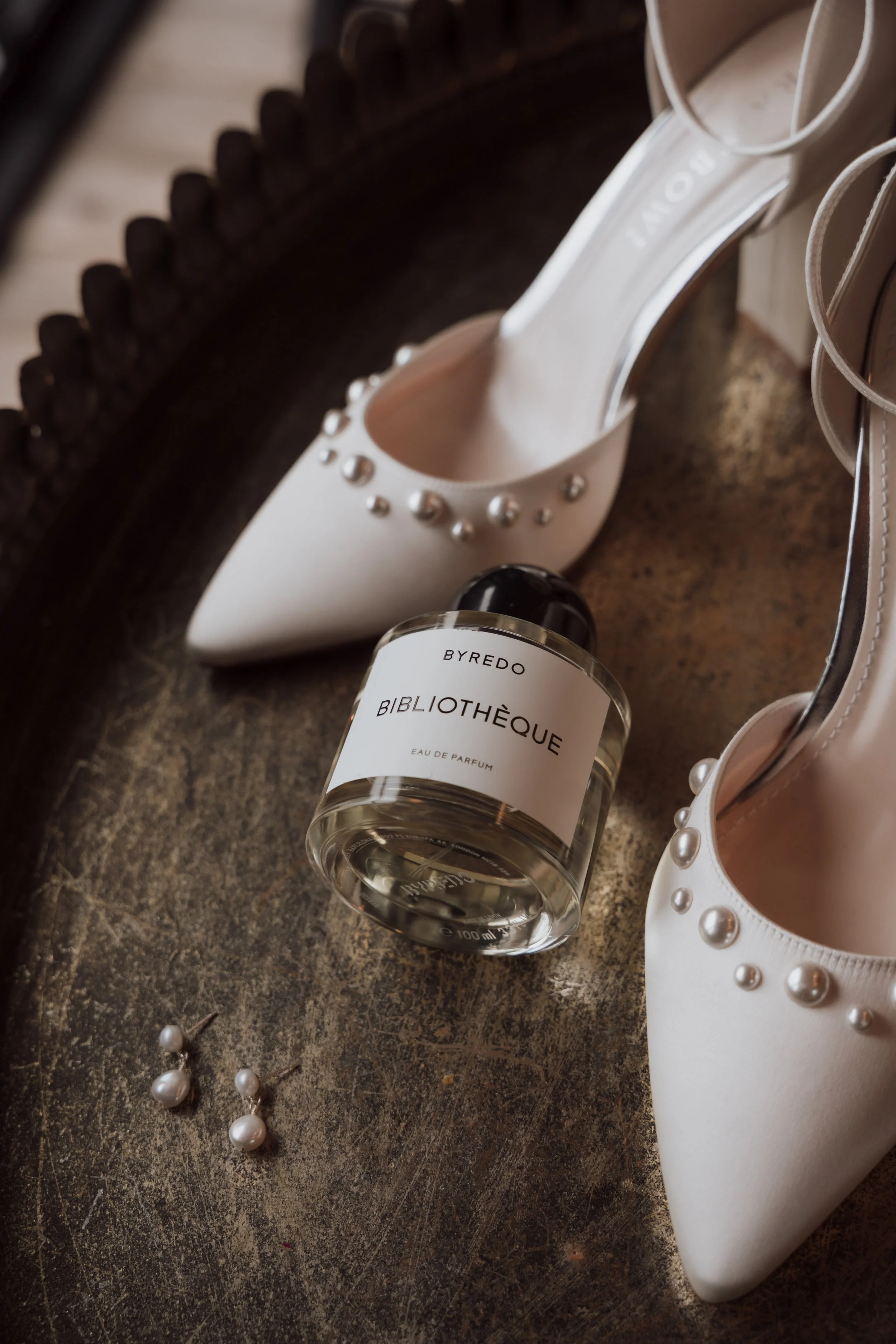 A pair of white high-heeled shoes with pearl embellishments, a bottle of Byredo Bibliothèque perfume, and a pearl necklace on a dark textured surface.