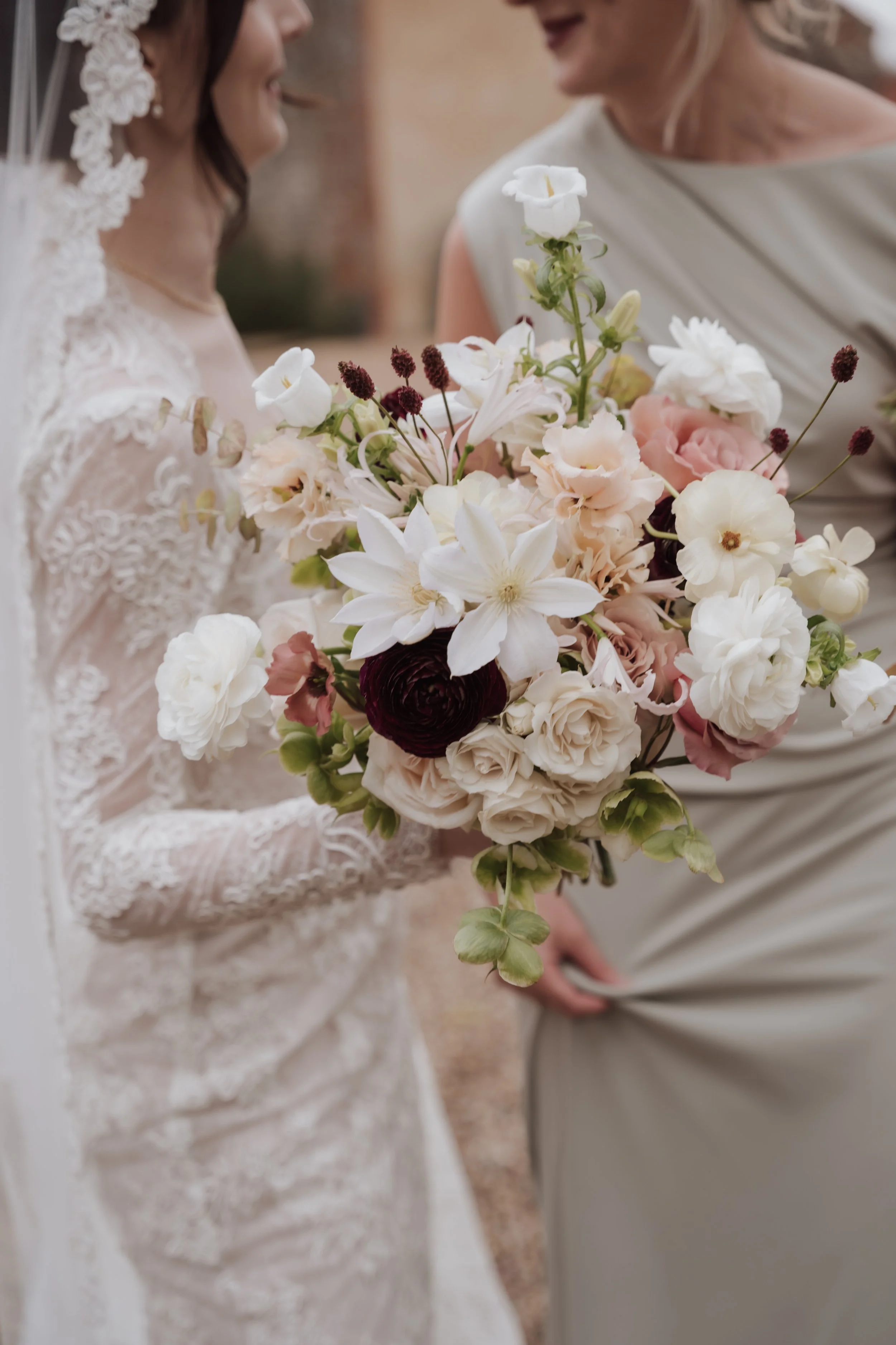 Close-up of a bride holding a bouquet of white, pink, and burgundy flowers, smiling with another woman in a light-colored dress during a wedding celebration.