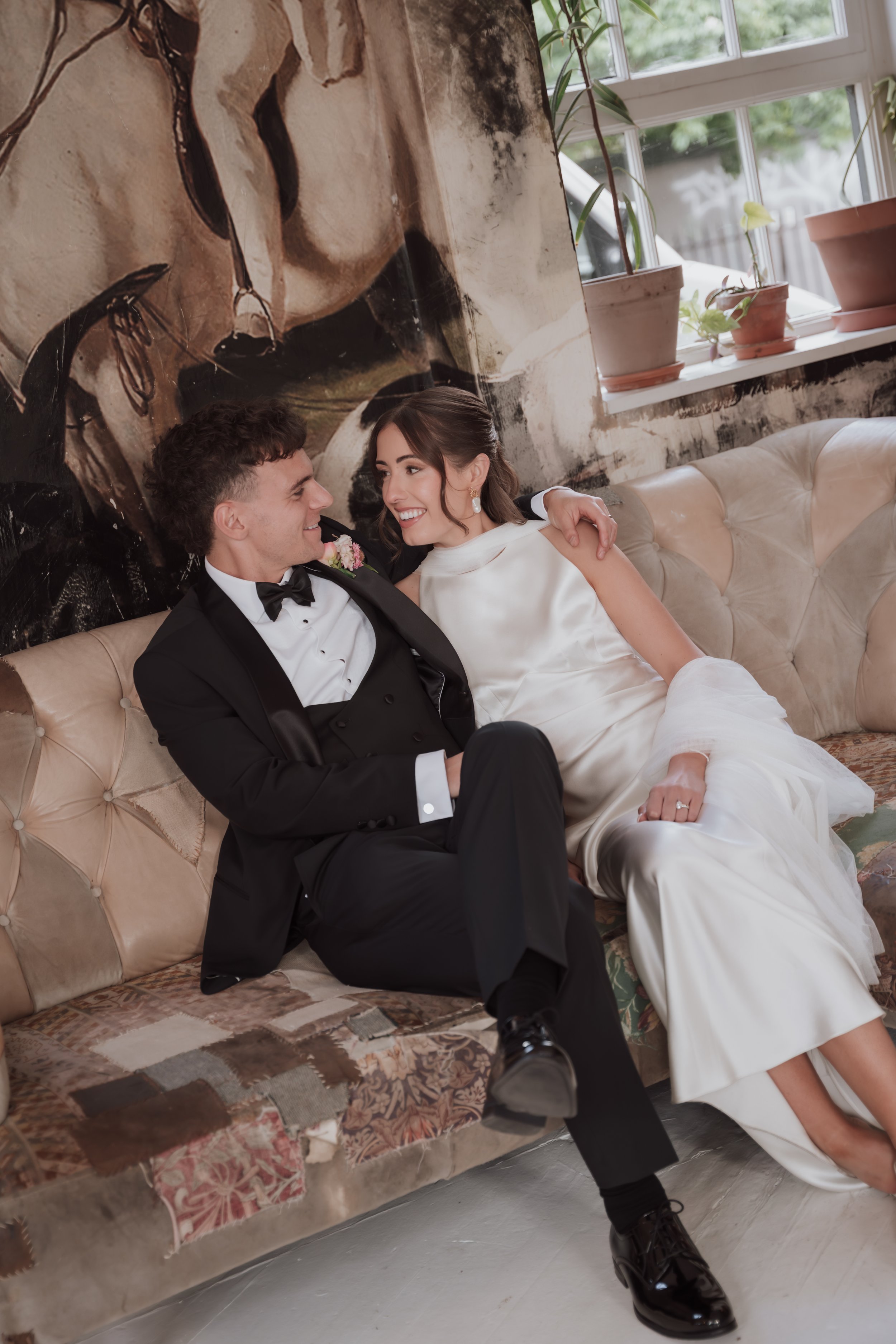 A bride and groom sitting on a vintage sofa, smiling and looking into each other's eyes indoors near a window with potted plants on the sill.