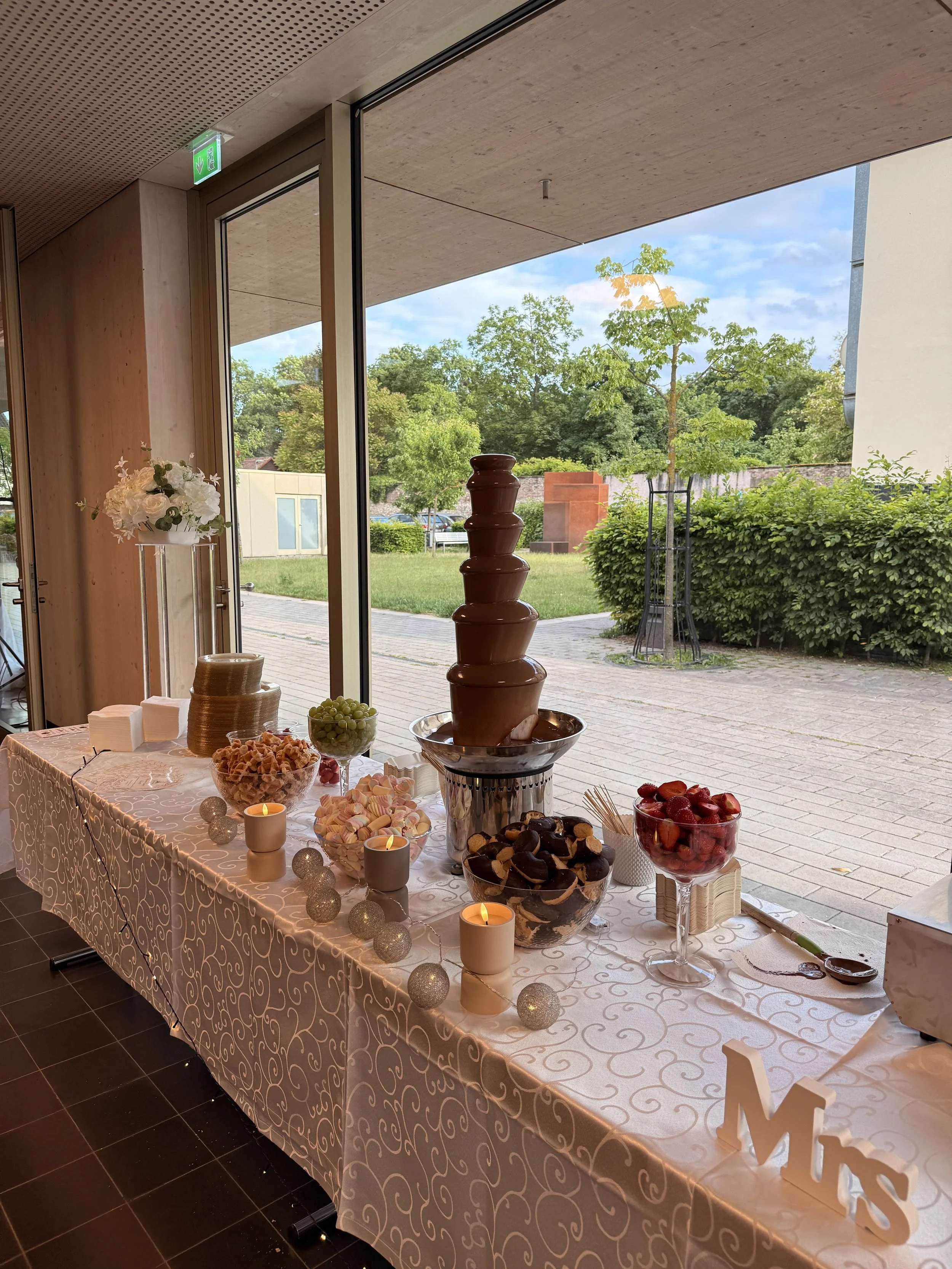 Ein Tisch mit Süßigkeiten, Kerzen und Dekorationen, vor einer großen Fensterfront mit Blick nach draußen auf Bäume und einen Weg.