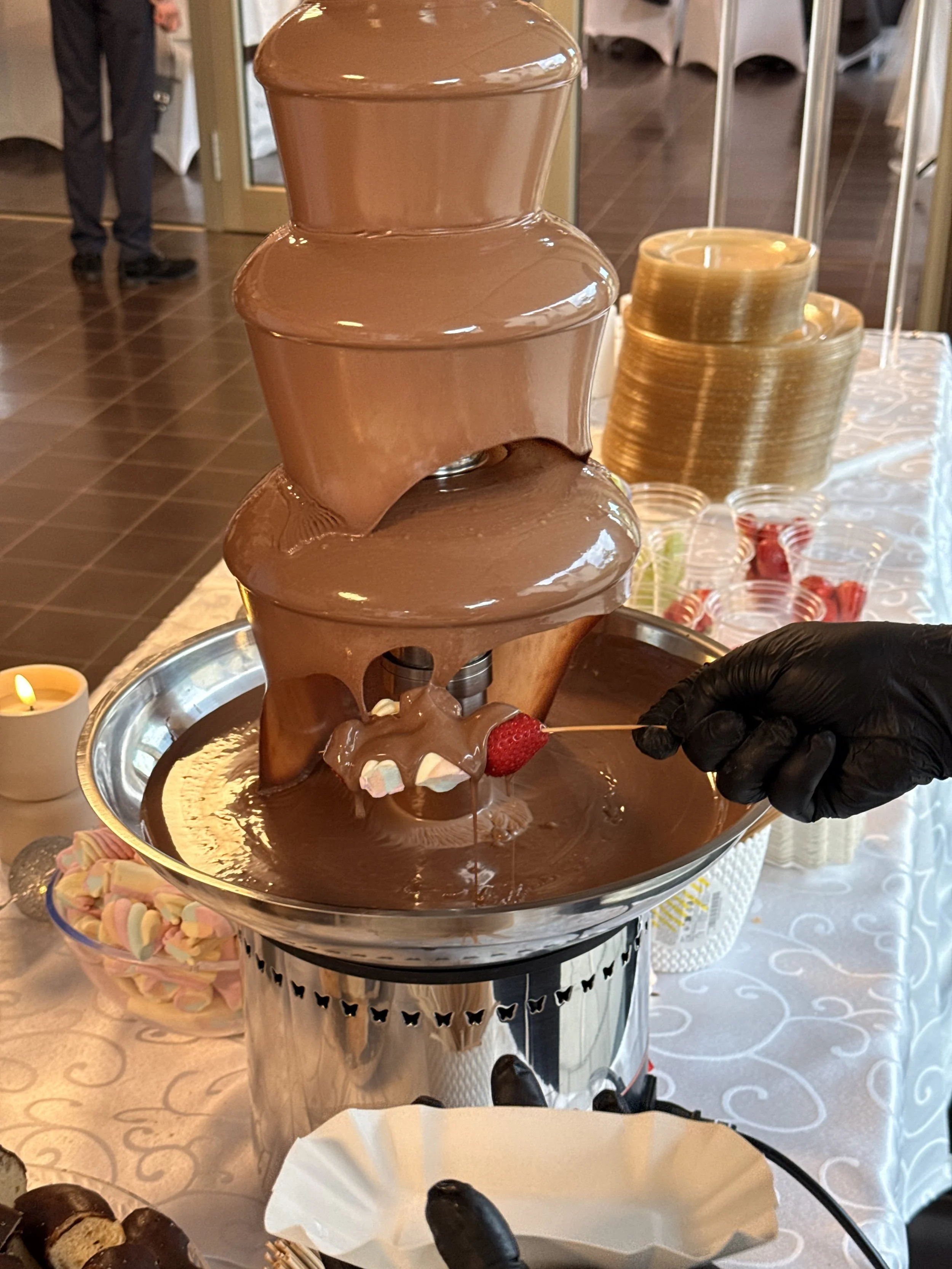 Ein Schokoladenbrunnen mit flüssiger Schokolade auf einer Hochzeitstafel, umgeben von kleinen Süßigkeiten und Obst zum Eintauchen.