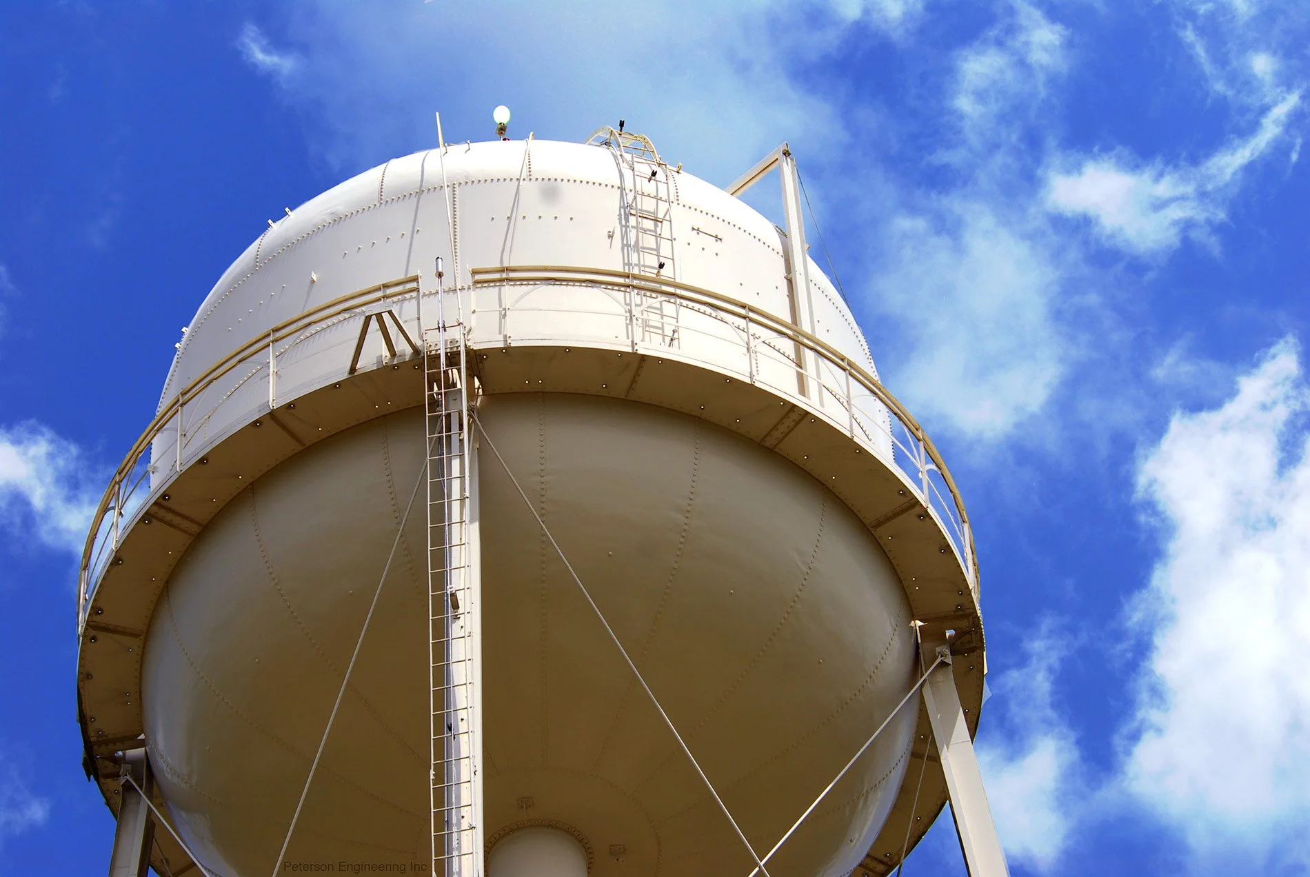 White water tower with a ladder and safety railing against a blue sky with some clouds.
