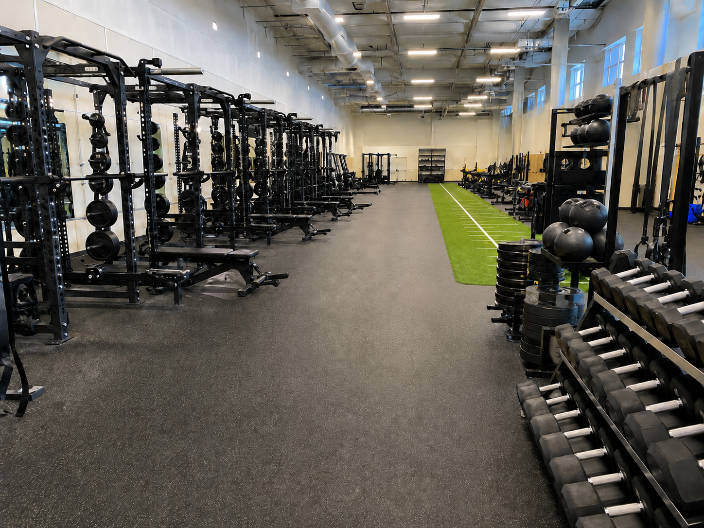 Modern college weight room with power racks, dumbbells, turf lane, medicine balls, and organized strength training equipment in a bright collegiate performance facility.