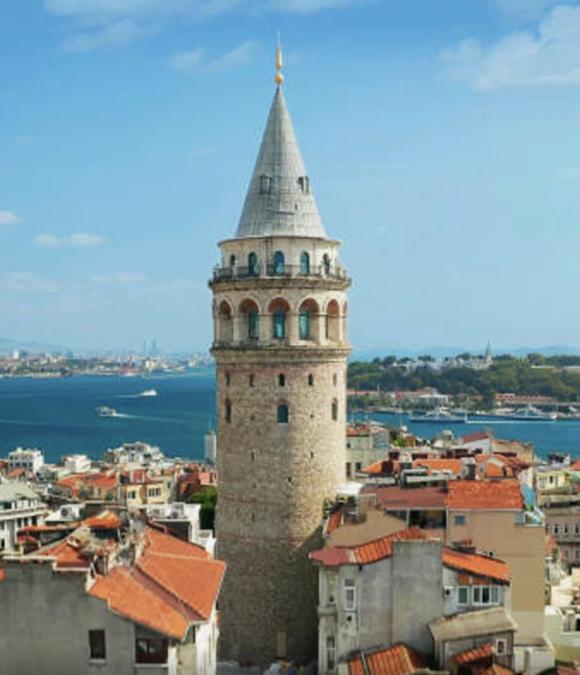Tall, stone tower with a conical silver-gray roof and narrow windows, surrounded by red-tiled roof buildings and overlooking a body of water with boats, under a partly cloudy sky.