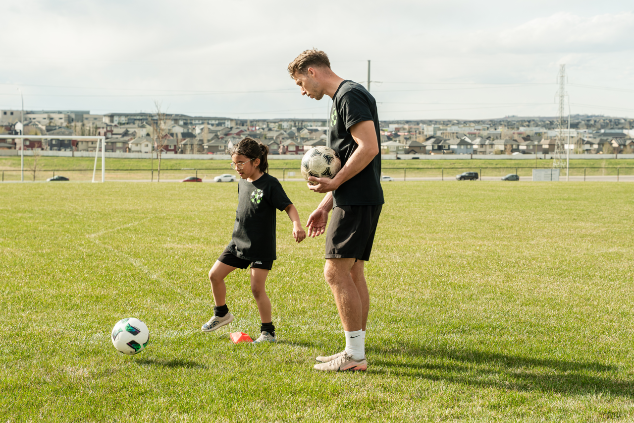 Soccer coach providing one-on-one technical training to a young player during a youth soccer skills session on a grass field in Calgary.