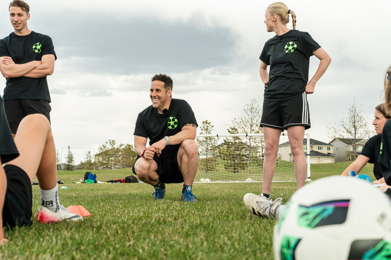A group of people in black sportswear gathered on a grassy field during daytime, some sitting and some standing, with a coach or trainer kneeling and smiling in the center, with a soccer ball in the foreground and a goal in the background.