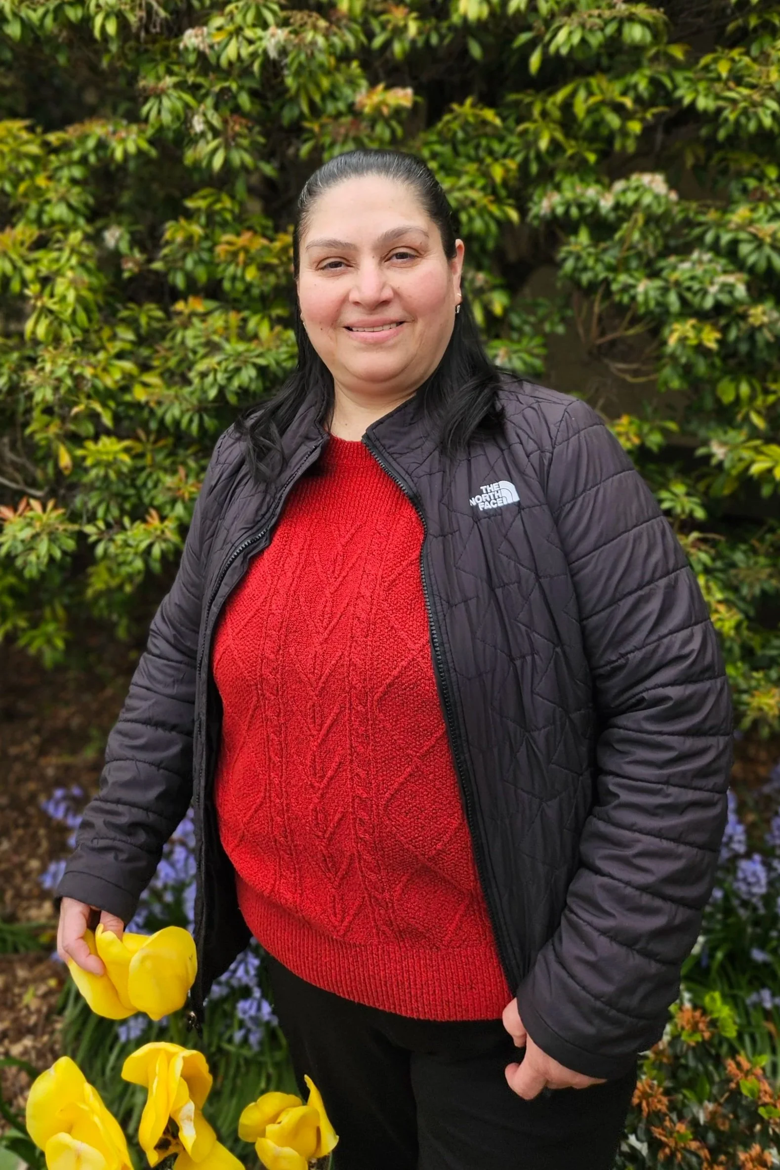 Smiling woman with dark hair pulled back, wearing a black North Face jacket and red sweater, holding yellow flowers, standing outdoors in front of green foliage with purple and orange flowers.