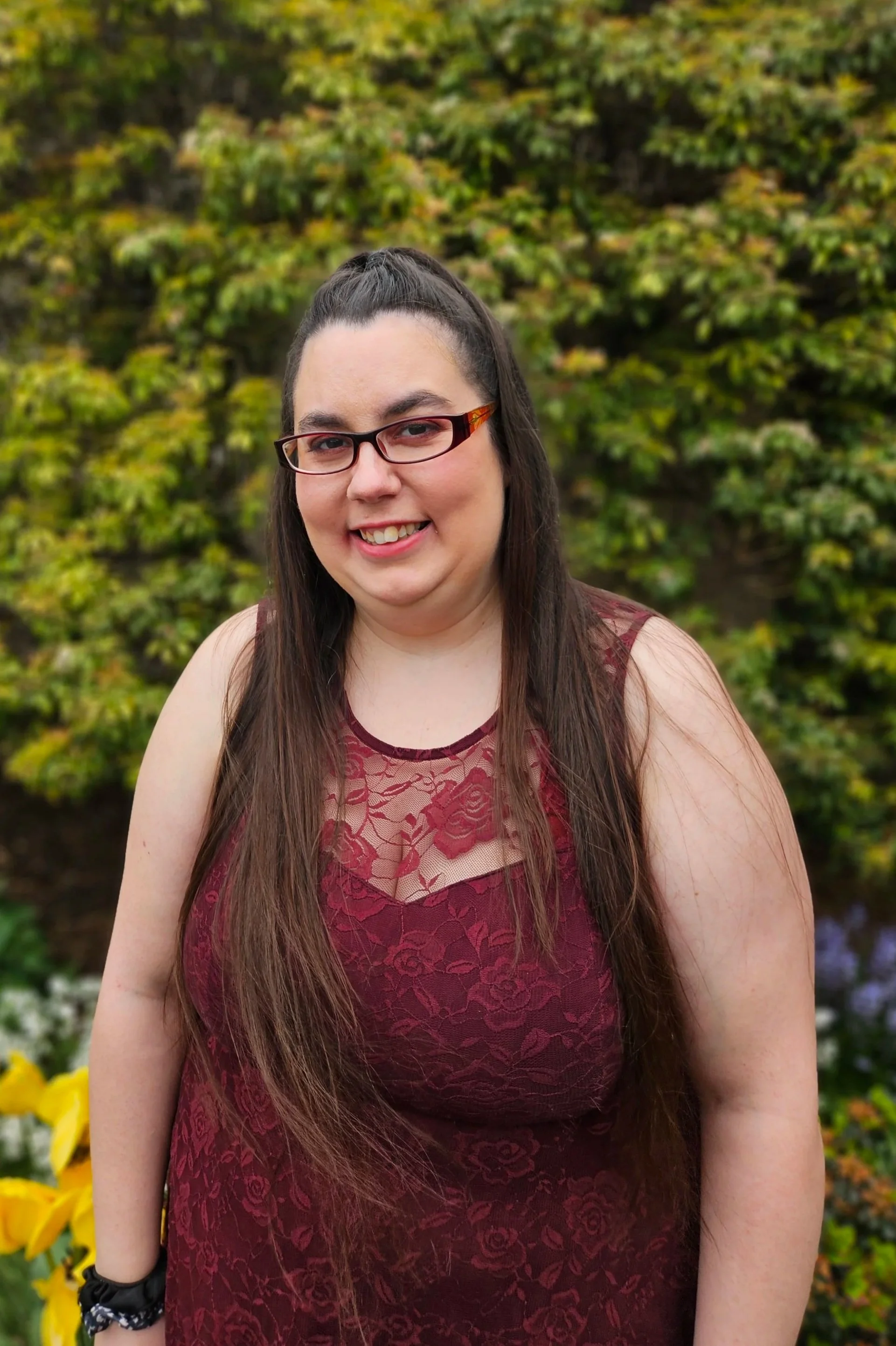 A woman with long brown hair, glasses, and a maroon lace dress, smiling outdoors with green and yellow foliage in the background.