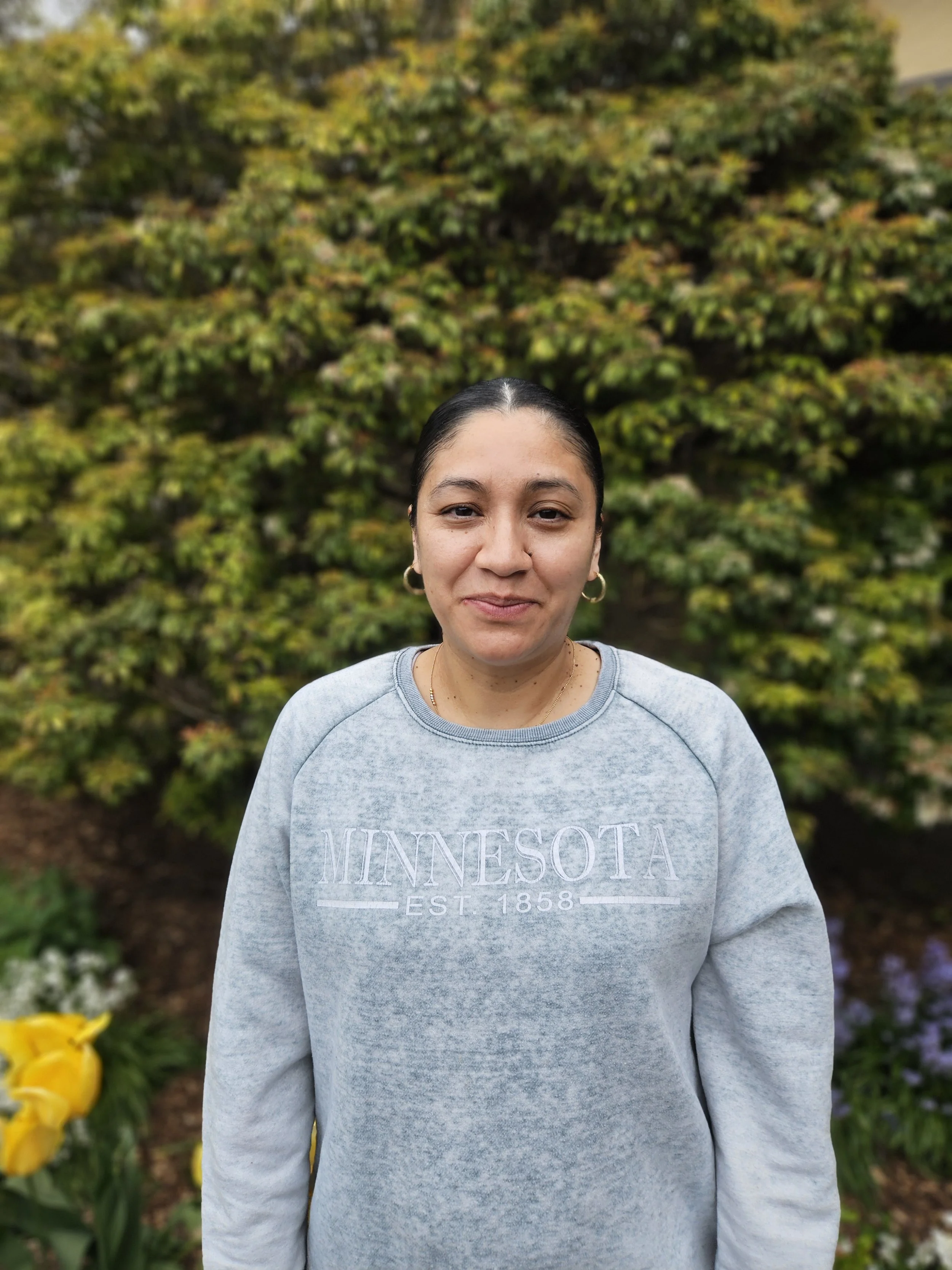 A woman wearing a gray Minnesota sweatshirt standing outdoors with lush green trees and colorful flowers in the background.