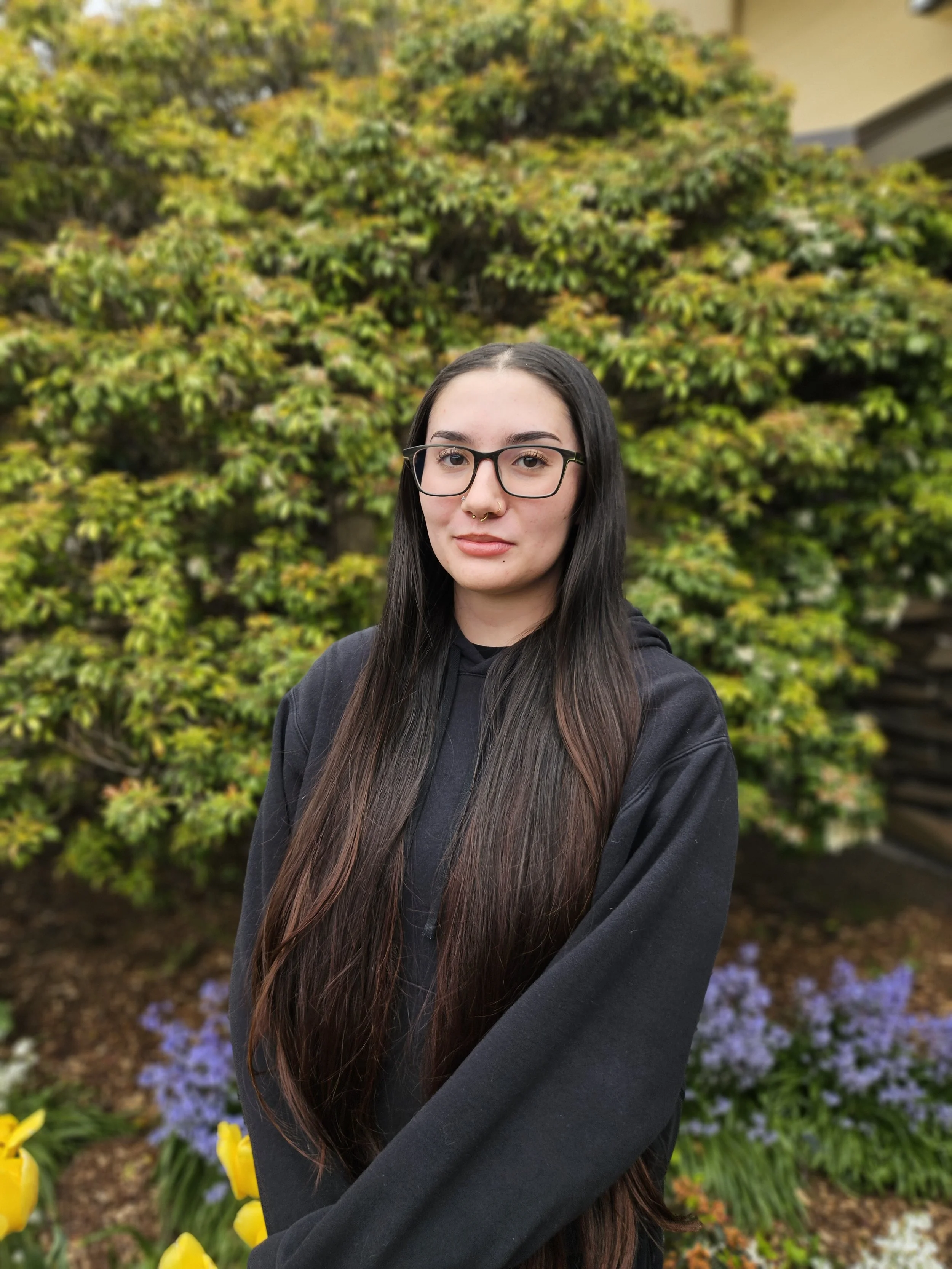 Young woman with long dark hair, glasses, and nose piercing, standing outdoors in front of a garden with yellow, green, and purple flowers.