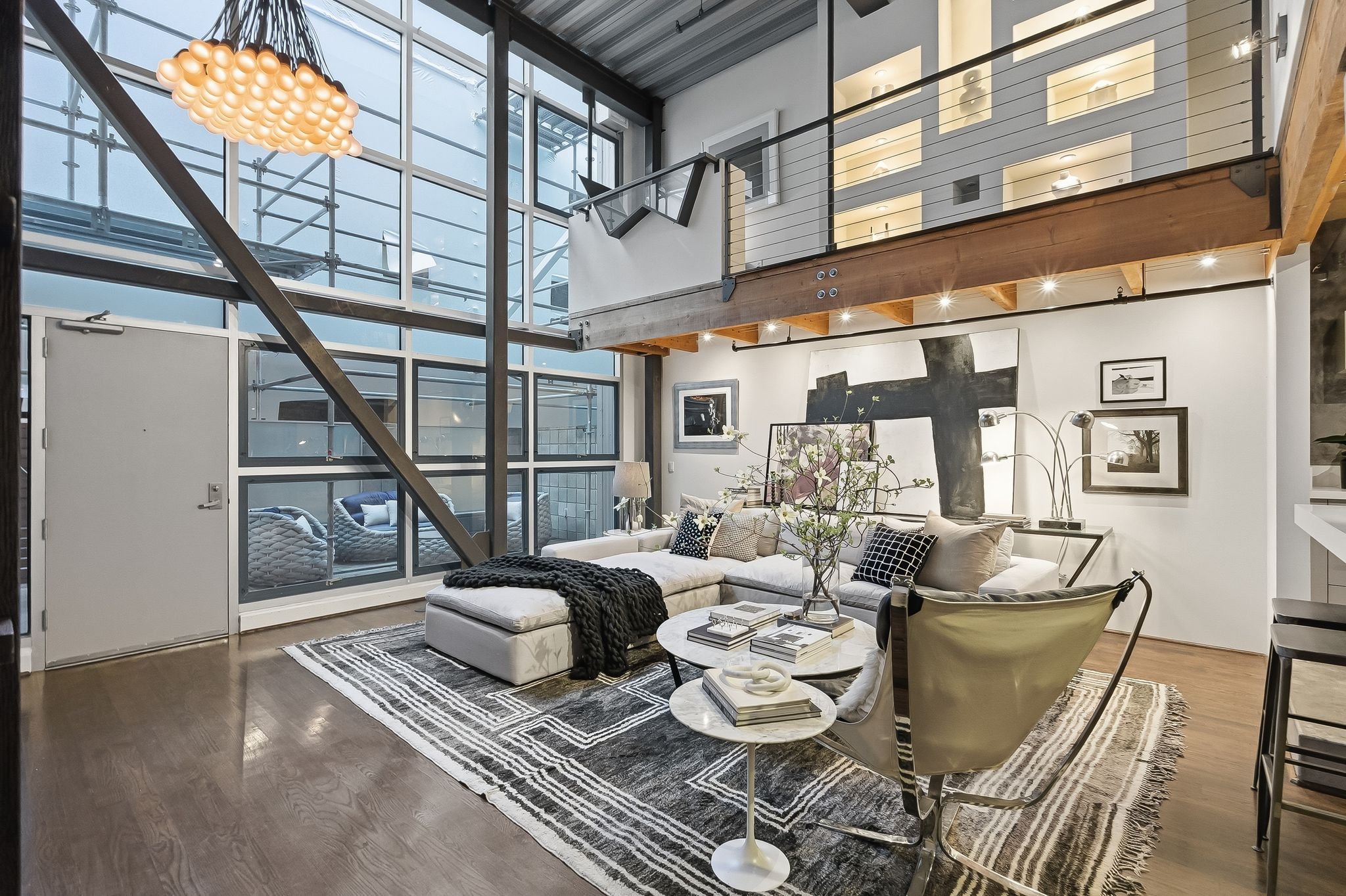 Modern living room with large windows, white sectional sofa, throw pillows, coffee table with books, lounge chair, abstract wall art, floral centerpiece, wooden floor, and a loft area with glass railing.